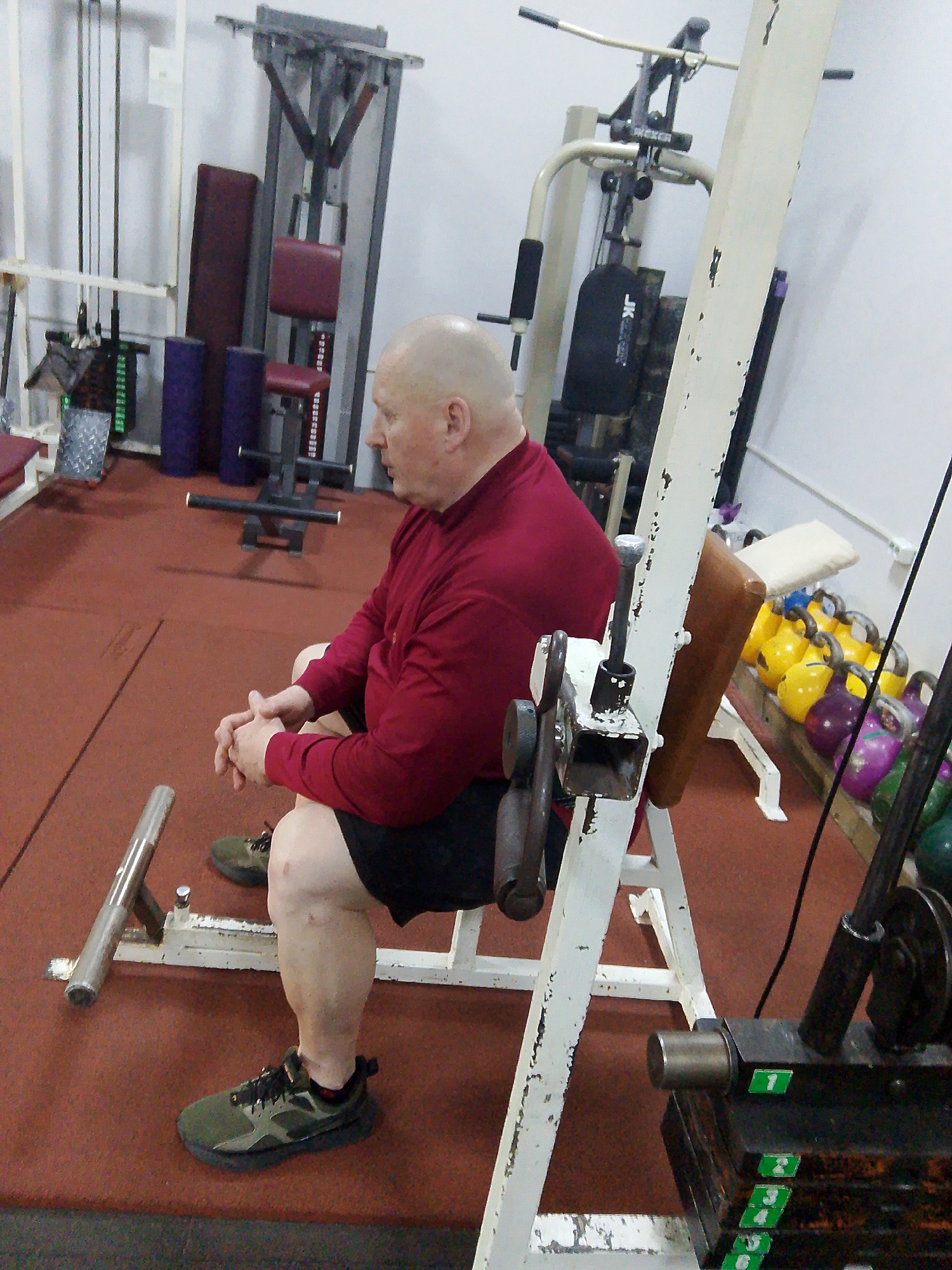 An older, bald man wearing a red long-sleeved shirt and black shorts is sitting on a weight machine in a gym. The man is facing to the left of the frame, with his hands clasped. He is wearing olive green athletic shoes. The weight machine is white and peeling, and has a brown pad behind the man's back. Weights with green number labels are stacked below the machine. In the background, other gym equipment is visible, including a cable machine, a lat pulldown machine, and a rack of kettlebells in various colors. The floor is a reddish-brown color. The location is a gym, possibly in the city of Chaikovsky, Russia.