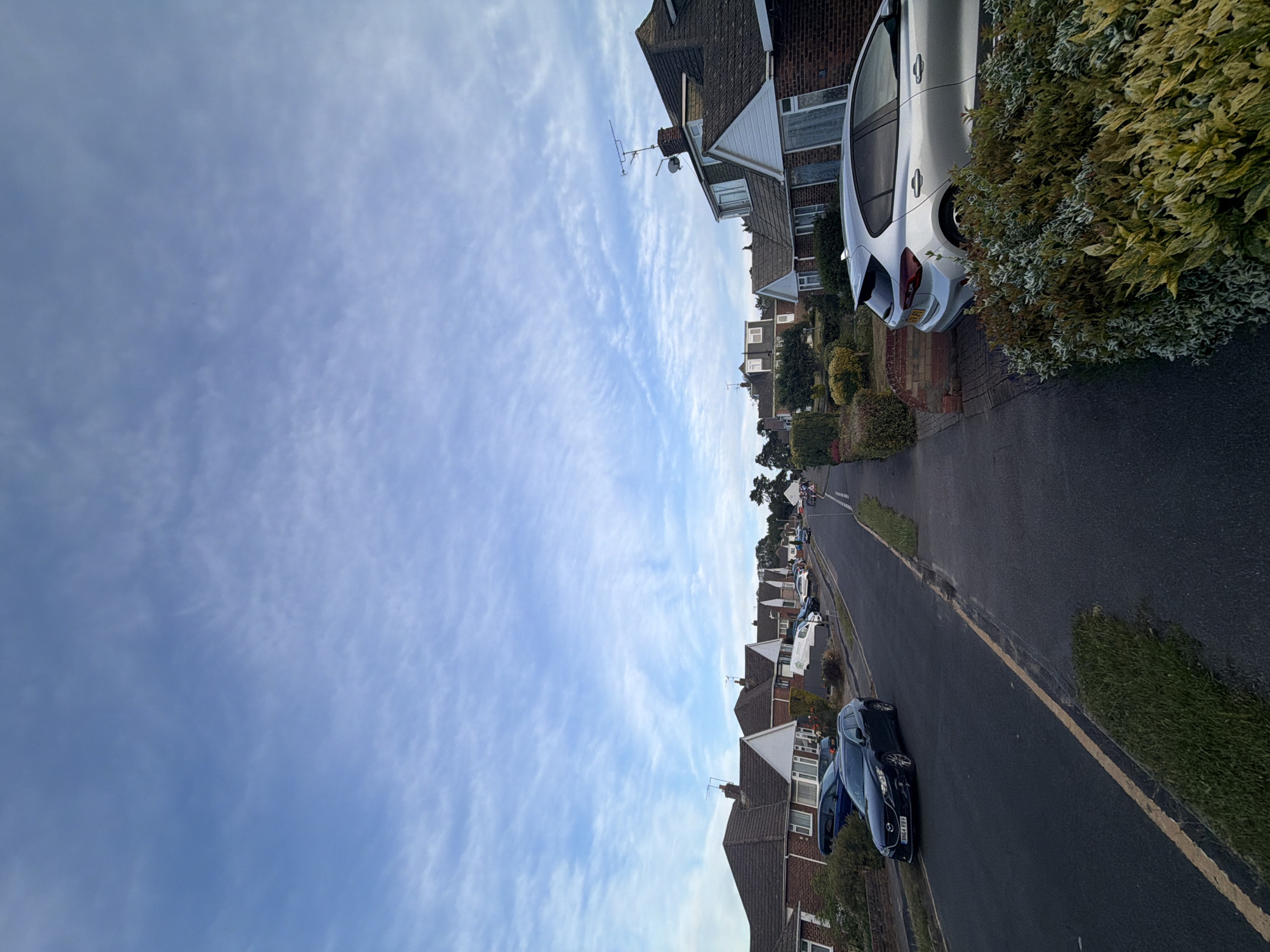 The image depicts a residential street scene in Maidstone, United Kingdom, viewed from a slightly elevated position looking up an incline. The sky dominates the upper portion, featuring a light blue hue with numerous thin, scattered cirrus clouds.
On the left side of the frame, a dark-colored Mazda 6 Estate (UK registration plate 'BN64 RBY') is parked partially on the curb and grass verge. Behind it, another dark vehicle is partially visible. Further up the street, a row of detached or semi-detached bungalows with brick facades, white gables, and brown tiled roofs line the road. Several other cars are parked along the left curb.
The asphalt road surface is dark grey, with a faded yellow line running parallel to the left curb, indicating parking restrictions.
On the right side of the frame, a dense, variegated hedge with green and yellow-green foliage is prominent in the foreground. Behind it, a silver Vauxhall Corsa hatchback (UK registration plate 'EY21 FJV') is parked on a paved driveway, its rear facing the viewer. Adjacent to this vehicle, a house with a brick ground floor, white upper gable, and brown tiled roof is visible. A chimney on this house is equipped with both a television aerial and a satellite dish.
Further along the street on the right, additional houses of similar architectural style are visible, bordered by hedges and small gardens. In the far distance, a small group of approximately three to four people can be observed walking on the right-hand pavement, near a white van. The street gently curves to the right as it ascends, and scattered trees are visible above the rooftops in the background.