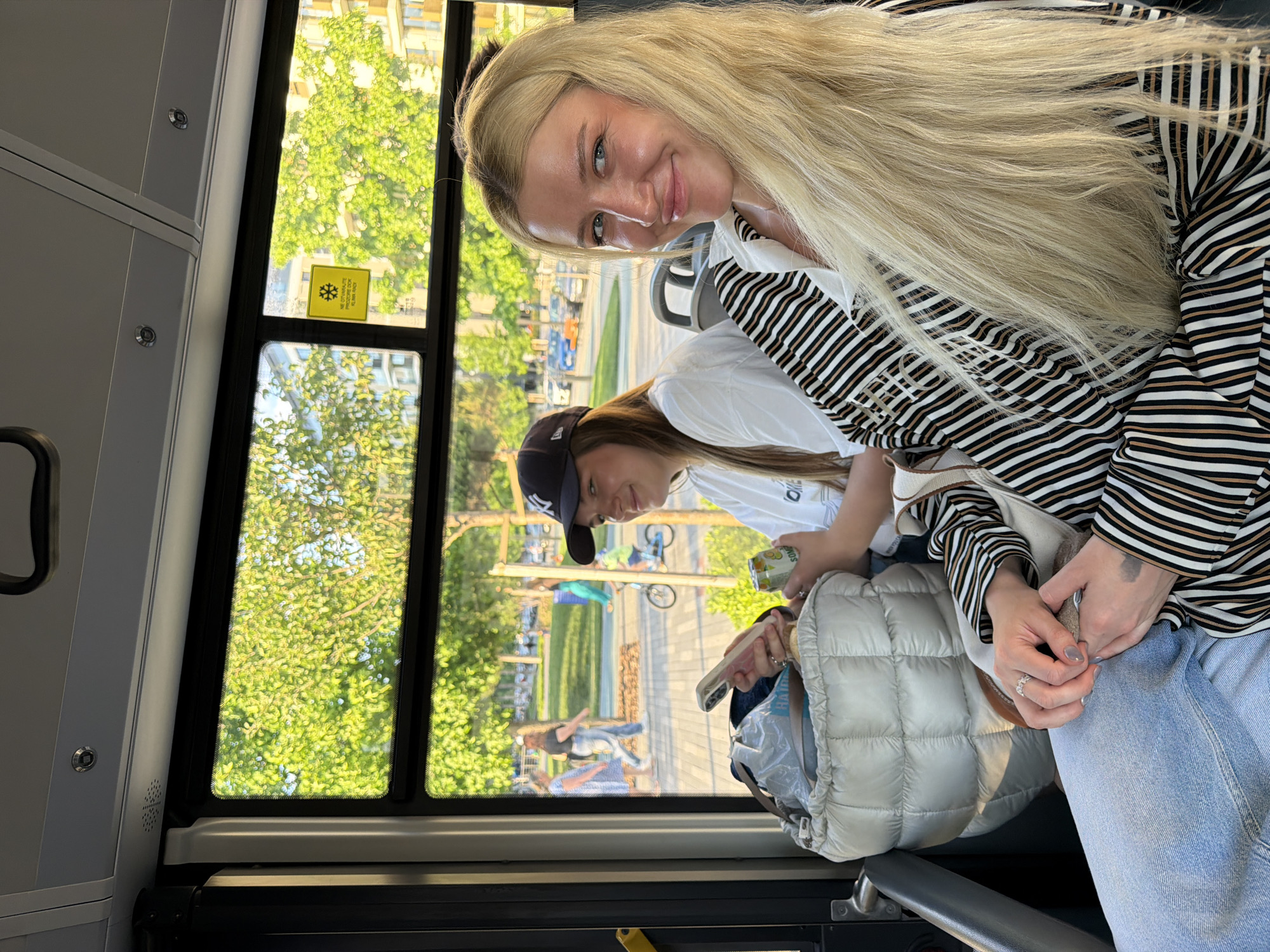 The image shows two young women inside a public transportation vehicle, likely a bus or train, as suggested by the window and interior paneling. The blonde woman, wearing a striped top and light blue jeans, is facing the camera and smiling. The other woman, wearing a white top and baseball cap, is looking down towards a bag on her lap. The reflection in the window reveals an outdoor scene with trees, a park area with people, and buildings in the background. Based on the yellow sticker on the window, the location is in Belgrade, Serbia.