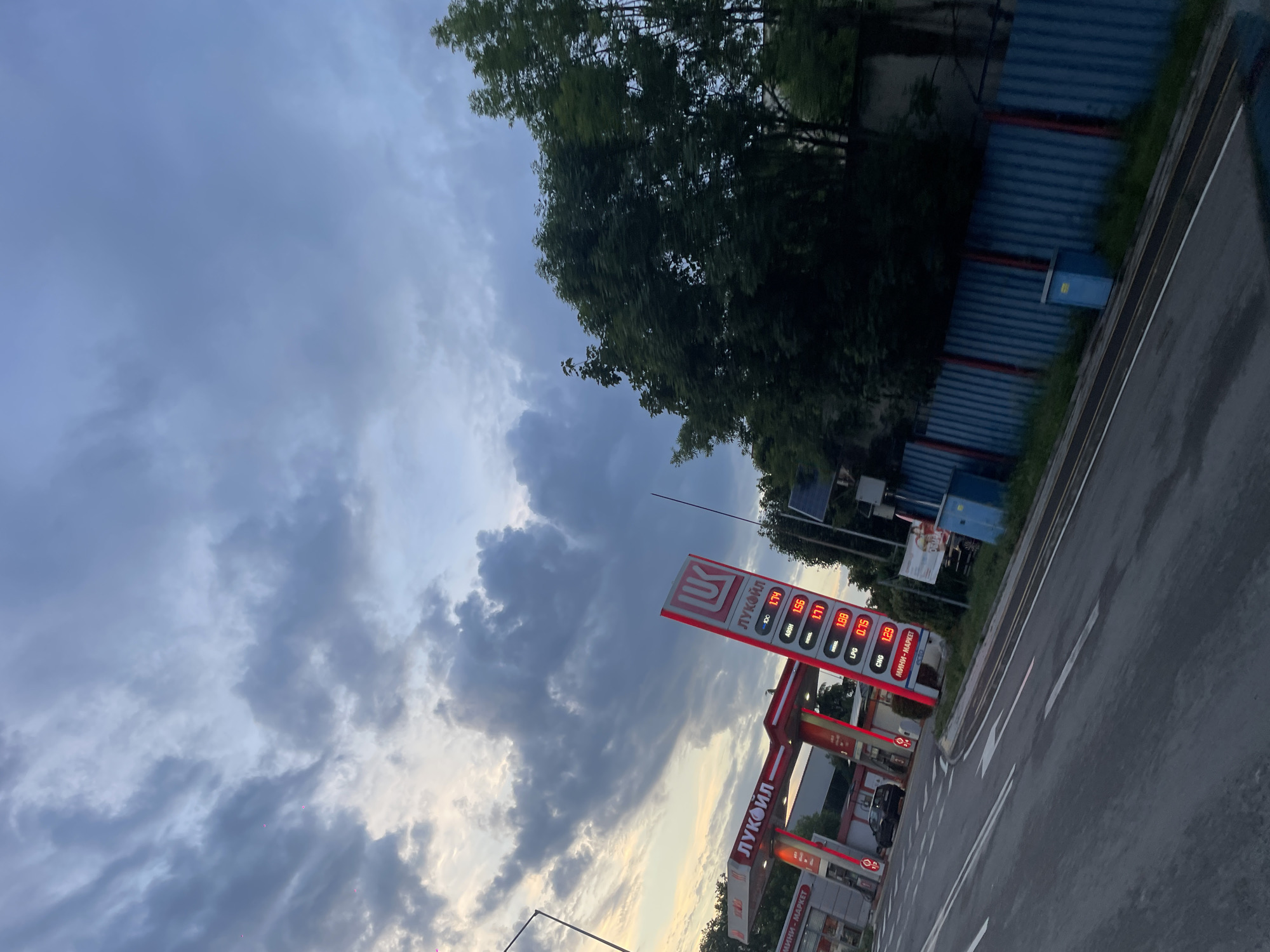The image captures a Lukoil gas station in Burgas, Bulgaria, under a cloudy sky. A tall price sign displays fuel costs, including 1.74 for 100, 1.56 for A95H, 1.71 for diesel, 1.88 for an unspecified fuel type, 0.75 for LPG, and 1.29 for CNG. The gas station's red and white branding is visible on the sign and the structure itself. The scene includes a glimpse of a road and nearby trees, with a blue corrugated fence in the background.