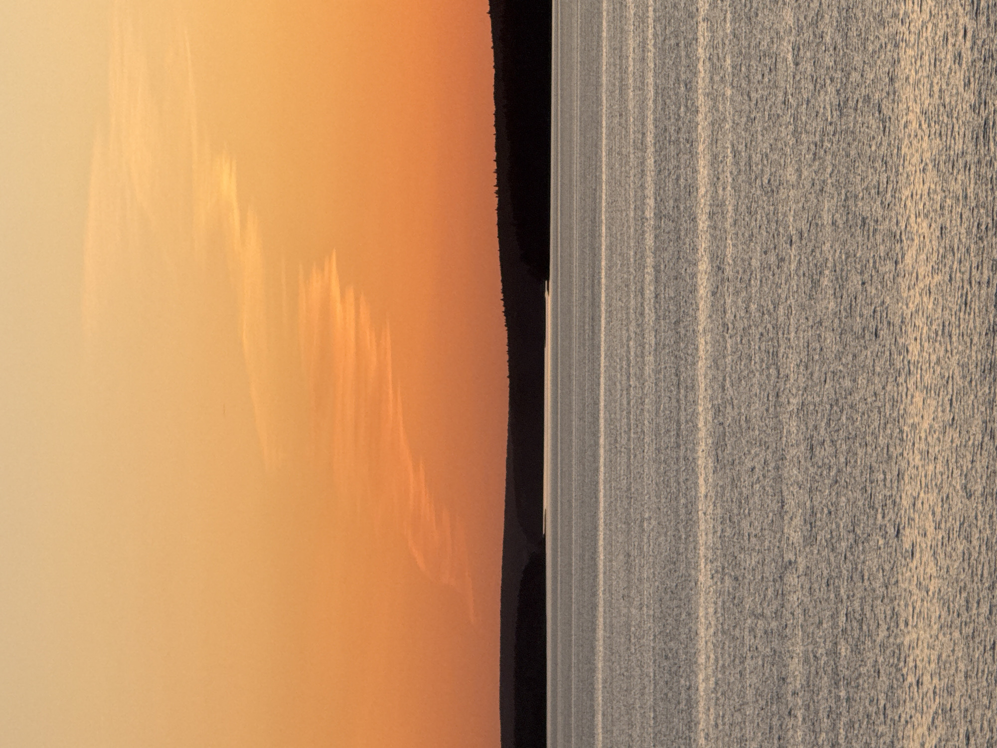 The image depicts a landscape with three distinct sections. On the left is a light orange sky with faint, striated cloud formations. In the middle is a dark, vertical strip that appears to be a wooded shoreline with trees. To the right is a body of water, potentially a river or lake, with a textured surface due to ripples or small waves. The scene evokes a sense of calm and stillness, possibly captured during sunrise or sunset. This does not give any indication of a location in the city of Votkinsk, Russia.