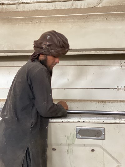 A man with a beard and turban, wearing dark clothing, stands next to a large, light-colored industrial machine, holding a metal bar. The environment appears to be an indoor industrial setting. There is no evidence to confirm the location is in Kandahar, Afghanistan.