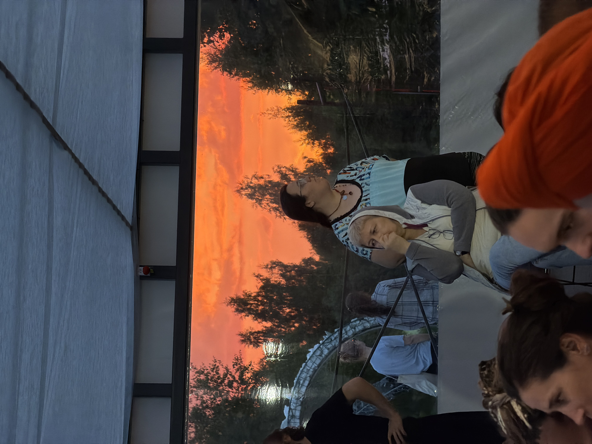 In Zhirnosovo, Russia, an outdoor gathering takes place beneath a large white tarp, casting a shadow over the scene. The backdrop is a large window or display showcasing a vibrant orange and red sunset over a dark forest. In the foreground, several people are visible, mostly from the chest up.
To the far left, a person in a black t-shirt stands with their back to the camera. Next to them, a woman in a light blue t-shirt and a necklace with beaded pendants is looking to the right, her hand near her face. In front of her, a person in a light gray hooded jacket and light blue jeans is seated, looking down with their hand still near their mouth. Behind them, a person in a light blue shirt stands with a white bag over their shoulder. Further back, another person in a plaid shirt is visible.
In the lower part of the image, several heads of attendees are visible, with one person in the far lower right wearing a bright orange head covering. The overall atmosphere appears to be one of quiet observation, perhaps at an event or performance, with the striking sunset providing a dramatic visual element.