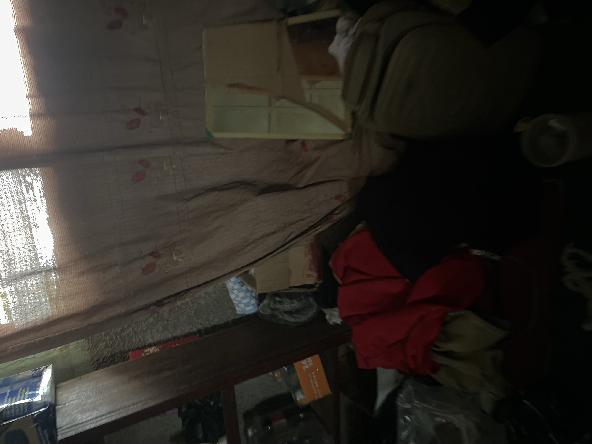 The image depicts a cluttered indoor space, likely a room, with various objects strewn about. A large piece of brown fabric, possibly a curtain or tarp, dominates the left side of the frame, featuring a floral pattern of red and yellow flowers. Light streams in from the left, indicating it is daytime. On the right side, a light-colored, somewhat bulky object could be a piece of furniture or stacked belongings, partially obscuring a dark, amorphous shape behind it. In the mid-ground, a small, open box-like structure with a light-colored frame is visible, appearing to be made of wood or particleboard and possibly containing shelves. Below this, a dark wooden surface, possibly a shelf or table, holds more items. A red garment, possibly a jacket or shirt, is folded or draped prominently on the right side of this surface, adjacent to other fabric items in lighter shades. Further down, a blue and white checkered item, perhaps a bag or piece of fabric, is visible. The overall impression is one of disarray and disorganization. There is no discernible activity taking place, and the lighting suggests it is daytime. No clear location cues are visible, and the emotions conveyed are neutral due to the lack of human presence or identifiable context. Some text might be present on packaging in the lower left, but it is too blurred to read.