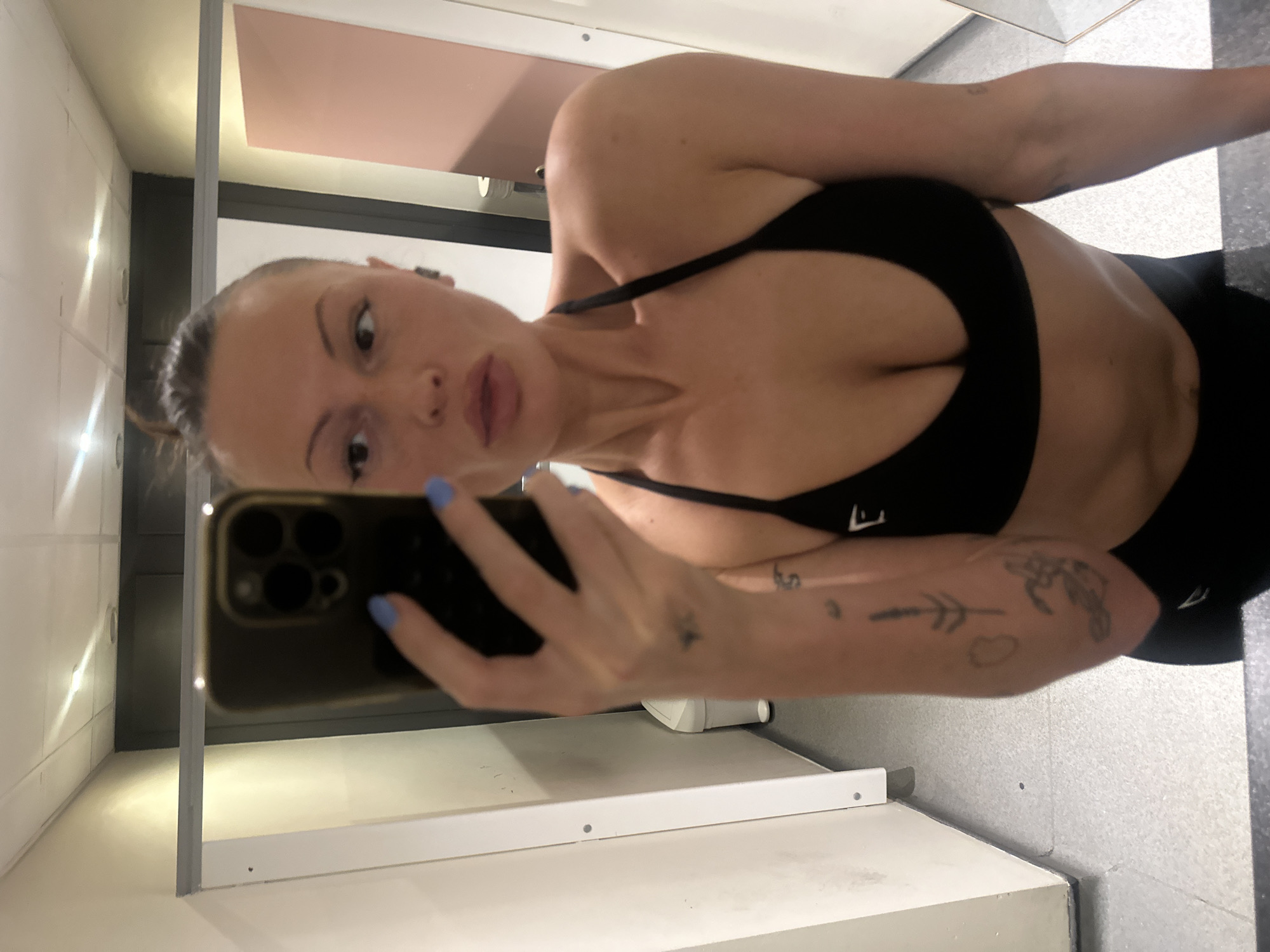 A woman is taking a selfie in a bathroom. She is wearing a black sports bra and black pants. Her hair is pulled back in a bun. She is holding a smartphone in her left hand. Her arm has multiple tattoos. The bathroom has a mirror and white countertops. The location is unknown and cannot be confidently determined as Tunbridge Wells, United Kingdom based on visual data.