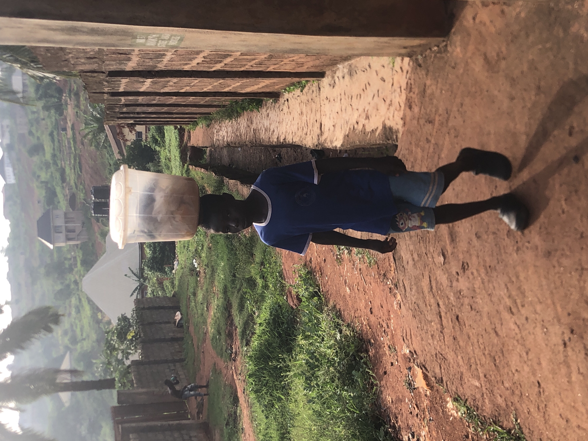 A young person is carrying a plastic bucket on their head. They are walking along a path. There is brick structure along the path, as well as foliage and trees. The location appears to be Ukwu-Abwa, Nigeria.