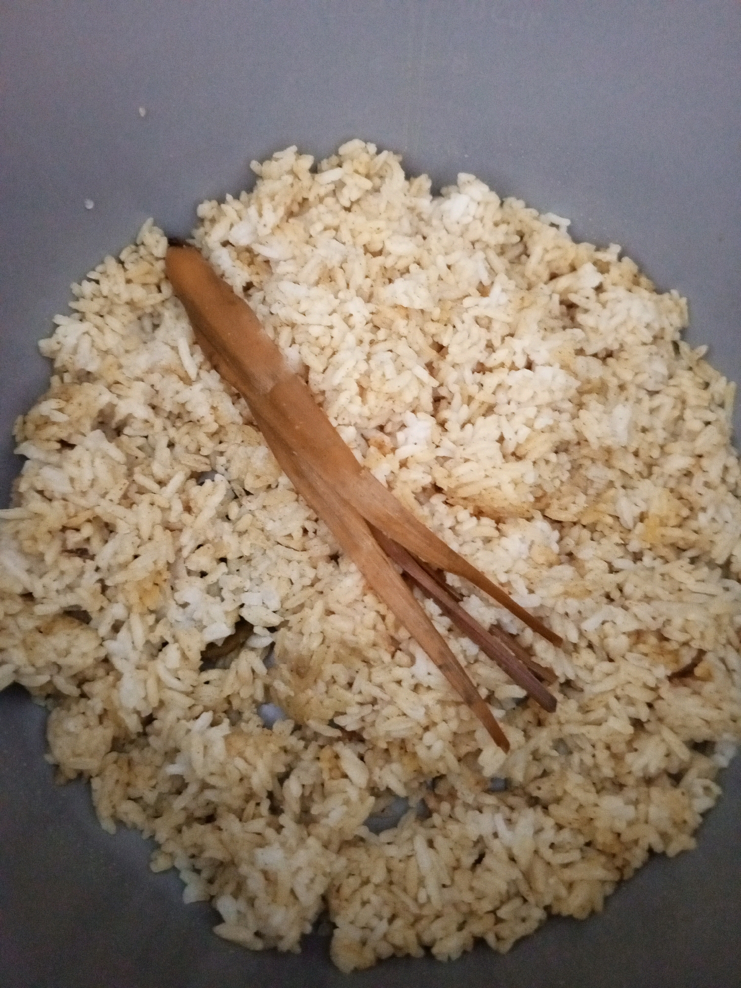 A close-up, overhead shot shows a pile of cooked brown rice in a grey pot. A wooden chopstick, split into three prongs at one end, rests on top of the rice. The rice appears slightly clumpy in places. There is no information in the image that indicates a specific location within Denpasar, Indonesia.