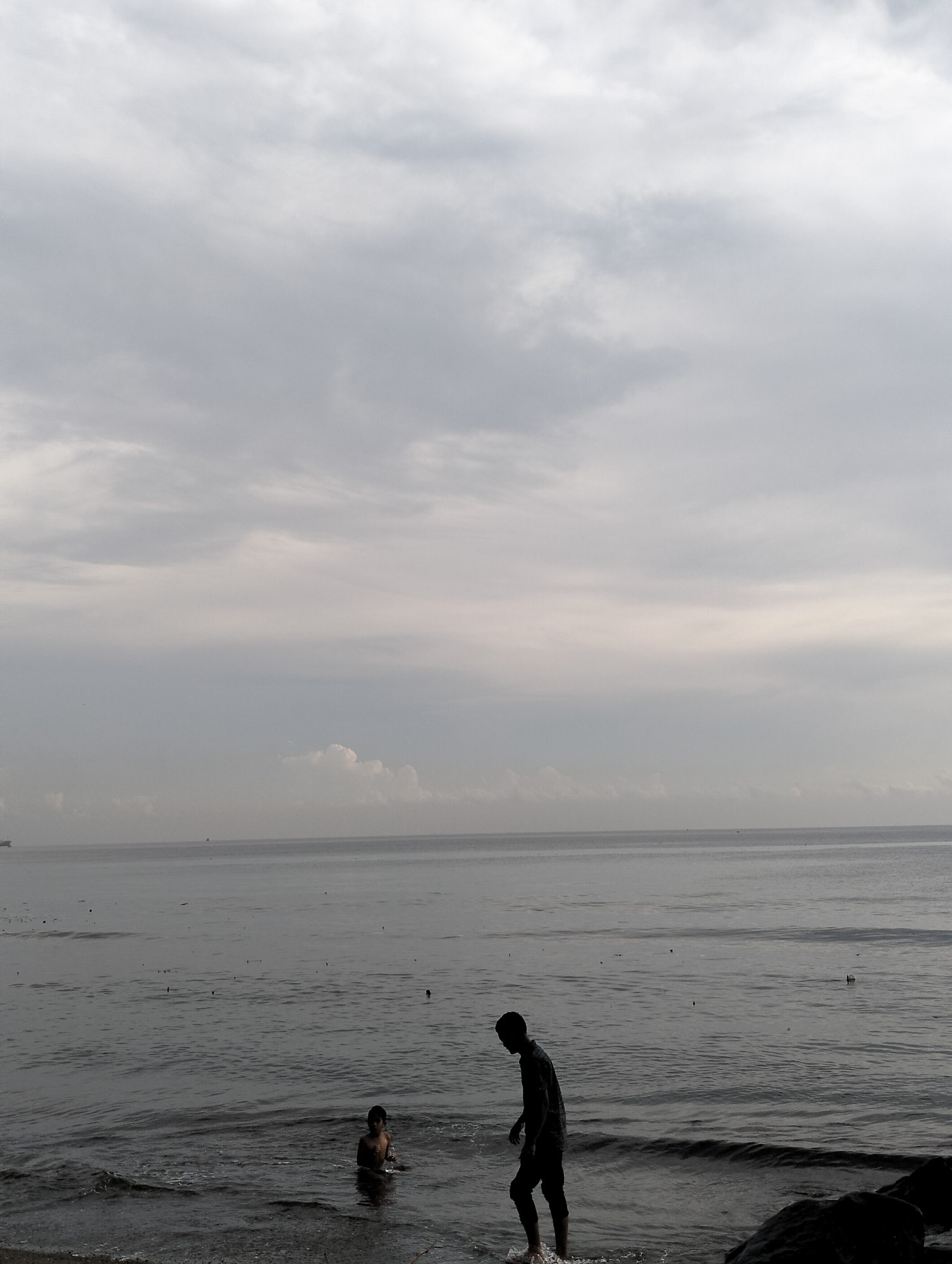 The image depicts a coastal scene in Lhokseumawe, Indonesia, under an overcast sky. The sea stretches to the horizon, with a faint outline of what appears to be a ship visible in the distance. Two individuals are present in the water near the shore. One person is standing in the water near some rocks. Another person is submerged to the mid-torso. Both individuals face away from the camera and are likely children or small adults. Debris is scattered on the water surface, including floating particles. The sky occupies the upper half of the frame and presents a uniform grey coloration. The overall scene is somber due to the muted color palette and overcast conditions.