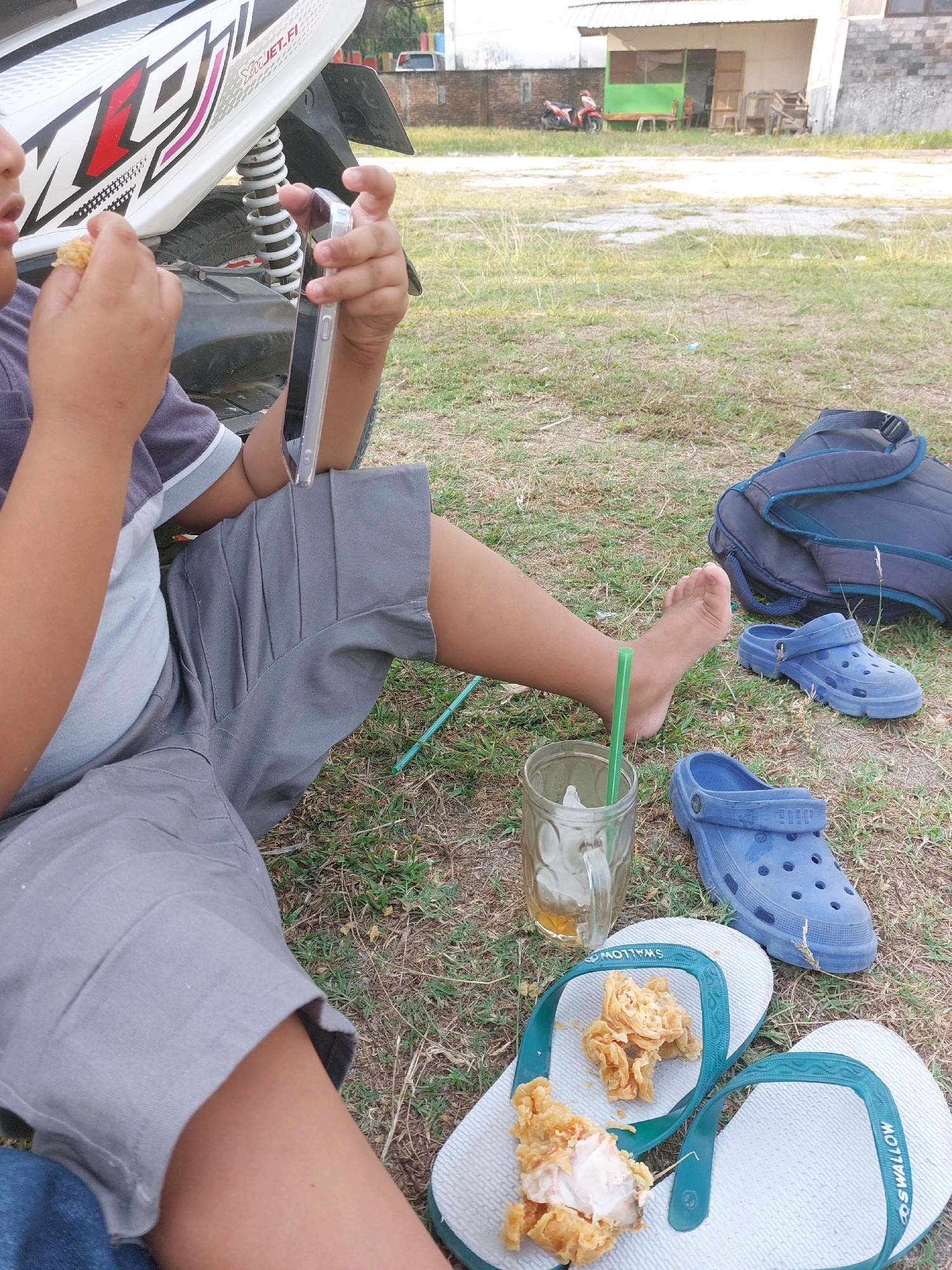 In the city of Sarangan, Indonesia, a young child, whose face is obscured, sits outdoors on grassy terrain next to a white motorcycle. The child is dressed in a grey and purple t-shirt and grey shorts. They are holding a smartphone in their right hand and a piece of fried chicken in their left. Nearby, a glass of iced tea with a green straw sits on the ground. A pair of blue clogs are placed next to the glass. To the right of the clogs, a navy blue backpack rests on the grass. A pair of white flip-flops, also on the ground, have pieces of fried chicken placed on them. In the background, a building with a corrugated metal roof and an open doorway is visible, with a red motorcycle parked in front of it. The overall scene suggests a casual outdoor break.