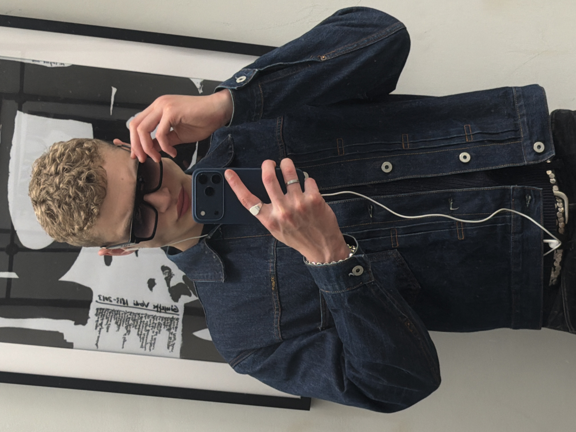 A person with short, curly blonde hair is taking a selfie in a mirror. The person is wearing sunglasses and a dark denim jacket over a dark top. The person is holding a blue smartphone in their right hand, with a silver ring visible on their ring finger. A white wired earphone is also visible. The mirror reflects a framed poster or artwork with black and white text and abstract images. The location is not identifiable as being in London, United Kingdom based on the image provided.
