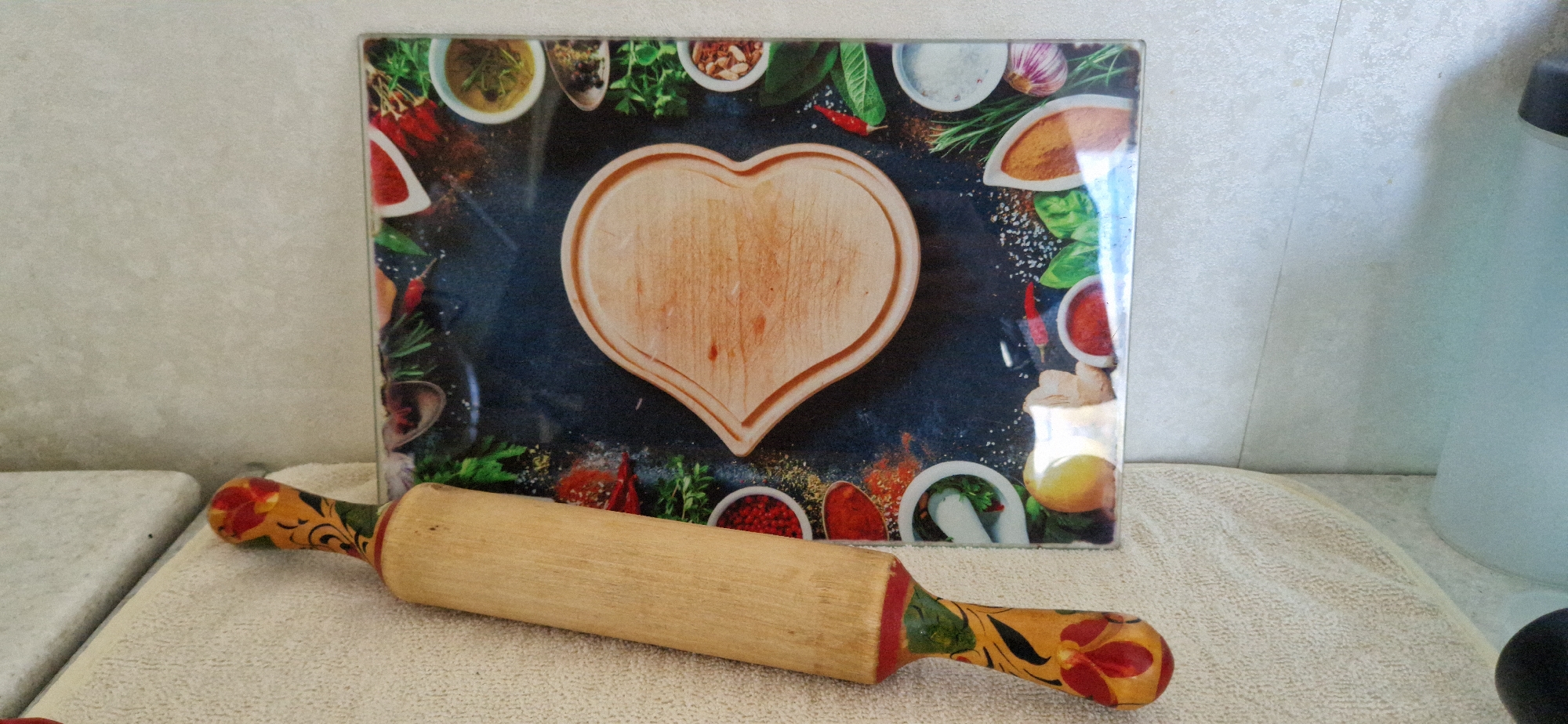 An overhead shot shows a wooden rolling pin with painted handles resting on a light-colored textured cloth. Behind the rolling pin is a rectangular glass cutting board with a dark background. The cutting board features an assortment of colorful spices, herbs, and sliced fruits arranged around a heart-shaped indentation in the center, which appears to be made of wood. The background of the image consists of a light-colored textured wall and a hint of a countertop.