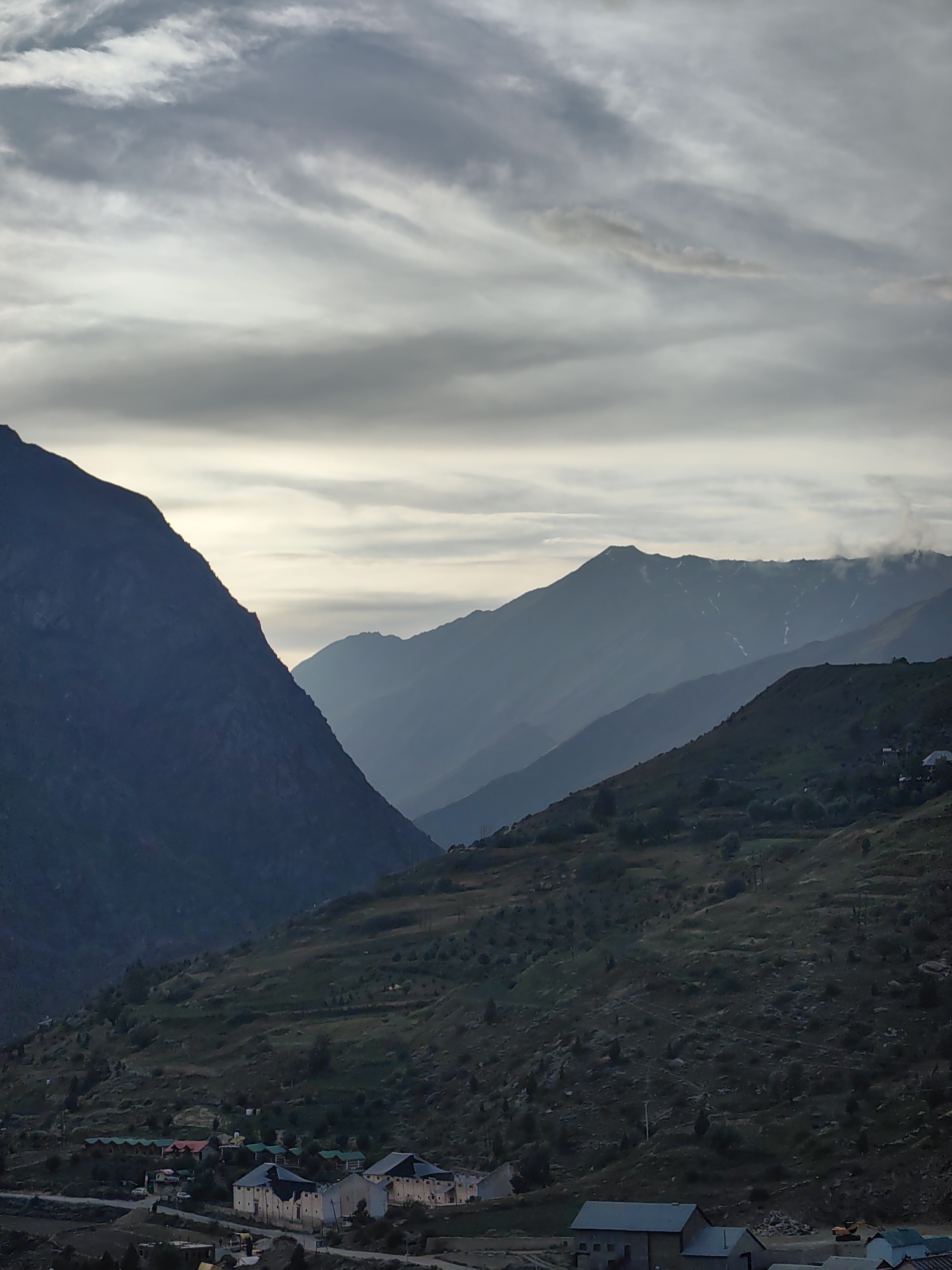 This is a photograph of a mountainous landscape with a village in the foreground, likely taken in Tandi, India. The sky is overcast with a gradient of grays, lighter near the horizon and darker above. Several layers of mountains are visible, fading into a hazy blue in the distance. The closest mountain on the left is dark and sharply defined, while subsequent mountain ranges appear softer and more muted due to atmospheric perspective. The slopes of the foreground mountains are covered in vegetation, with a scattering of rocks and trees. A village is situated at the base of the slopes, featuring buildings with a variety of roof colors, including green, red, and dark gray. Some of these buildings appear to be part of a resort or guesthouse complex, with a row of smaller structures with green roofs lining a road. Larger buildings with gray roofs and visible windows are also present. The overall lighting suggests it may be early morning or late afternoon, given the soft, diffused light. There are no people visible in the image.