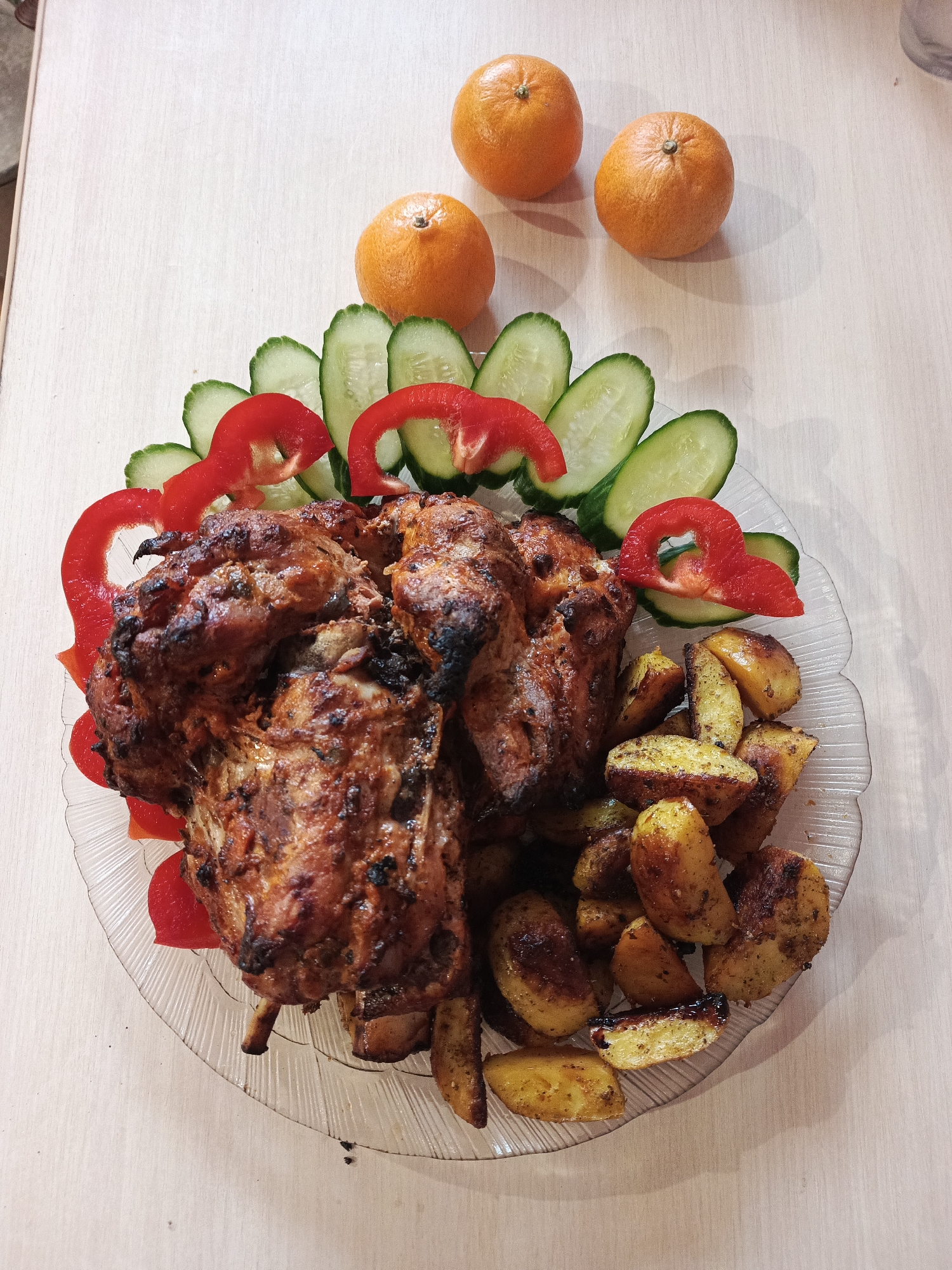 The image shows a prepared meal on a light-colored table. The meal consists of roasted meat, sliced cucumbers, red bell pepper rings, and roasted potatoes, all arranged on a clear glass plate with a scalloped edge. Three tangerines are placed above the plate. The photo was taken in Chaikovsky, Russia.