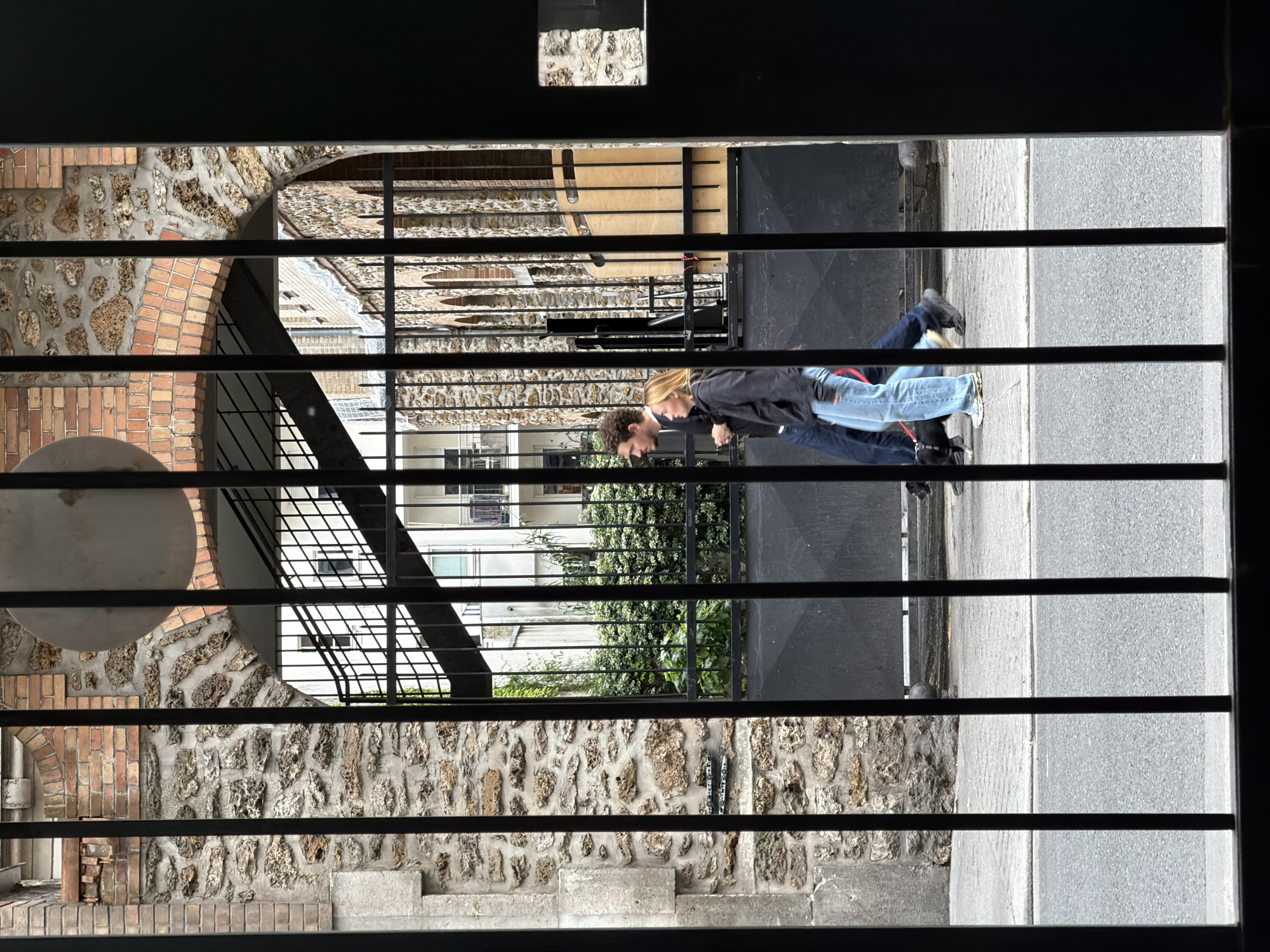 The image shows a couple walking side-by-side on a sidewalk, viewed through a gated entryway. The man has dark hair and wears a dark jacket and jeans. The woman has blonde hair and wears a dark jacket and light-colored jeans. To the left of the sidewalk and the gated entryway, a building with a mix of brick and stone construction is visible. In the background, beyond the gate, there are other buildings, some with green foliage. The image appears to be taken in an urban environment, potentially in Paris, France, given the architectural style.