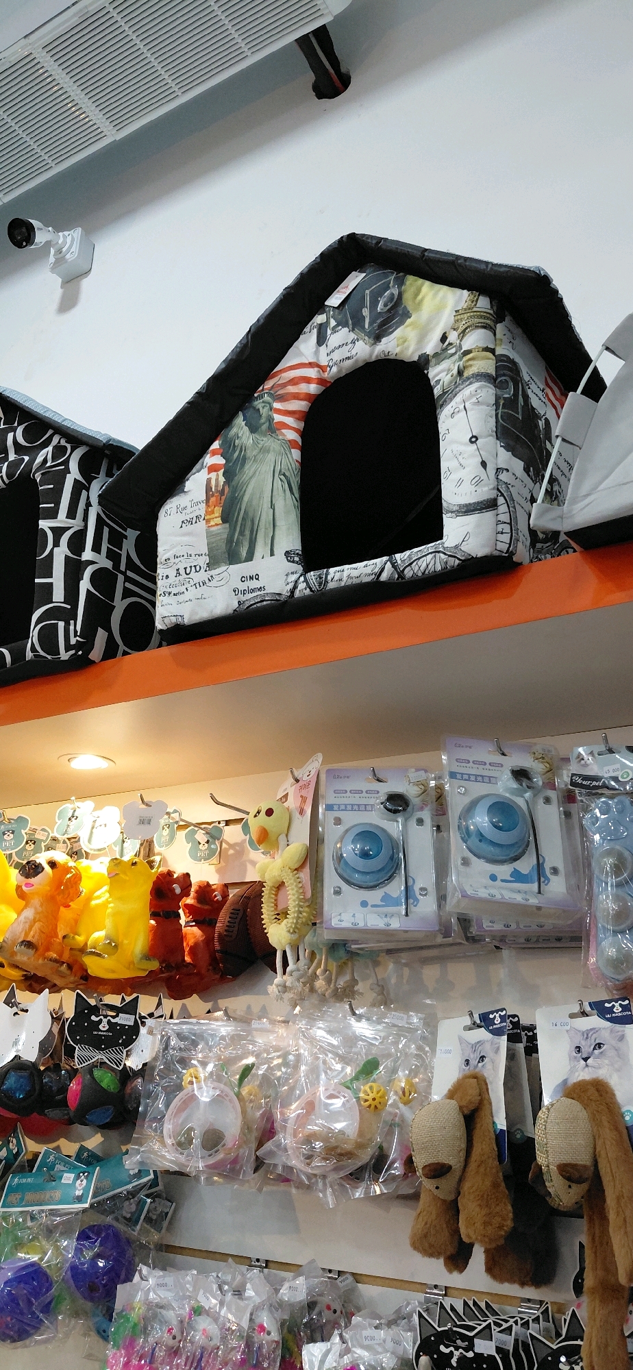 The image shows a retail store display featuring various pet products. In the foreground, shelves are stocked with colorful toys, including stuffed animals that resemble dogs, rabbits, and footballs, as well as a blue plastic ball. There are also packaged pet toys and accessories, such as interactive treat dispensers and small plush cat toys. In the upper portion of the image, two fabric pet houses are displayed, one with a black and white pattern and the other decorated with images of the Statue of Liberty and French text. A white security camera is visible on the wall above the pet houses. The overall environment appears to be a pet supply store, possibly located in Encarnacion, Paraguay, as indicated by the presence of currency denominations like "9.000" and "16.000" on some product tags, which are consistent with Paraguayan currency (Guaraní). There are no people visible in the image.