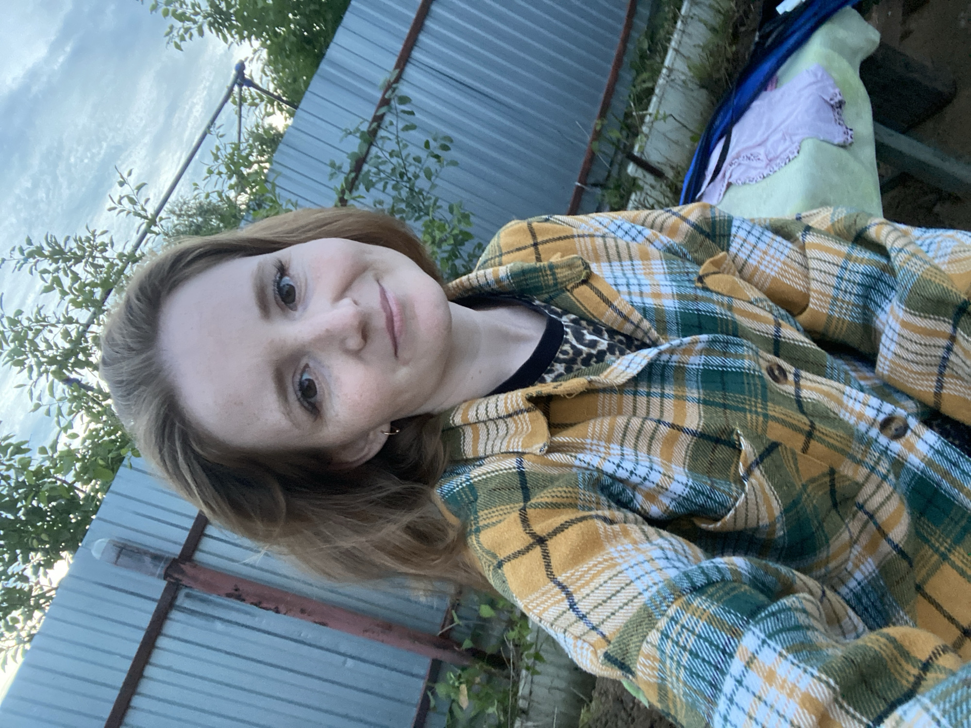 The media file shows a young woman taking a selfie in an outdoor setting. She has light brown hair, fair skin with freckles, and brown eyes. She is wearing a yellow and green plaid shirt over a leopard-print top. Her expression is neutral with a slight smile. The background suggests a rural or semi-rural location with a corrugated metal fence, some greenery, and a cloudy sky. The overcast sky indicates it might be daytime, possibly late afternoon or early evening. There is no visible text in the image. The presence of the corrugated metal fence and the general outdoor environment are consistent with a location in Chaikovsky, Russia.