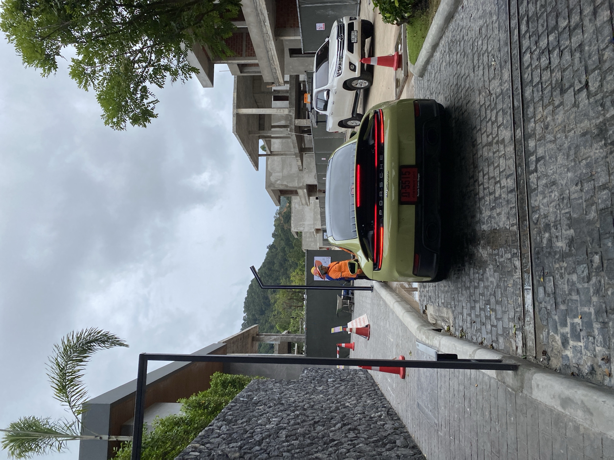 The image shows a partially constructed building site in Choeng Thale, Thailand, under an overcast sky. A green Porsche Taycan is parked on a cobblestone road with its taillights visible. Behind it, a white pickup truck is parked near orange traffic cones. An individual wearing an orange vest and hard hat stands near the Porsche. The building is multi-story and appears to be in the early stages of construction. A stone wall and foliage are visible in the foreground. A black metal frame structure is also present in the lower left corner of the frame.