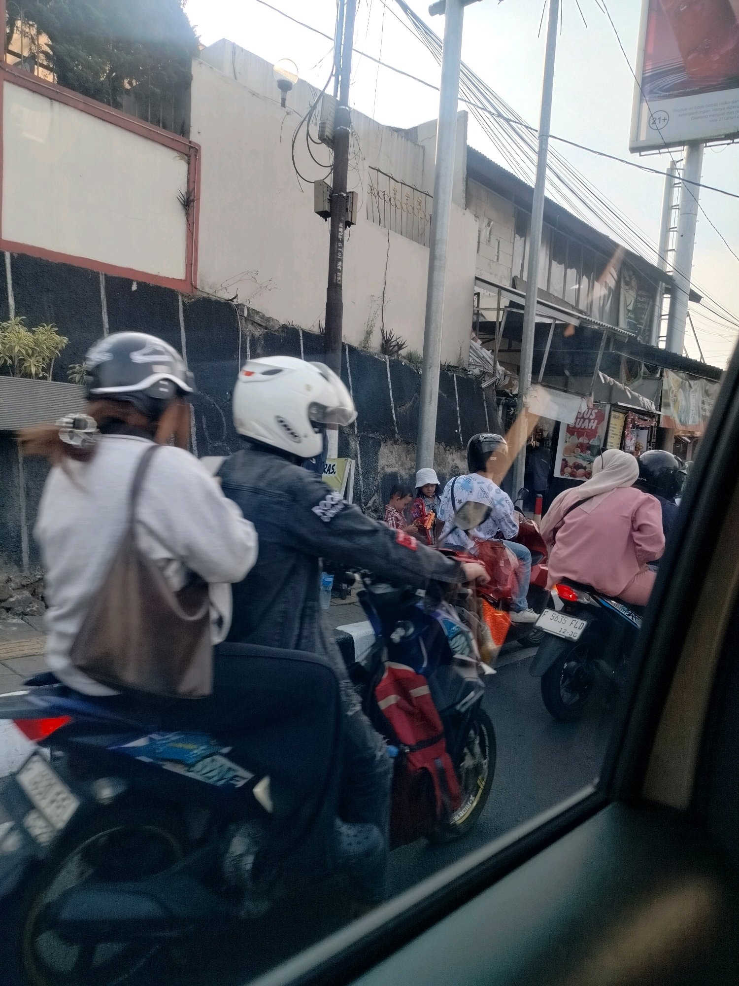 The image depicts a street scene in Cisarua, Indonesia. Several individuals are riding on motorcycles in what appears to be traffic. On the left, a person wearing a grey helmet and a white long-sleeved shirt is riding on the back of a motorcycle, with their hair tied up. In front of them, another individual in a dark denim jacket and a white helmet is driving. To the right, a person in a pink jacket and a light-colored hijab is on the back of a motorcycle, with the driver wearing a black helmet. Further back, other motorcycles and pedestrians are visible. The environment includes buildings, streetlights, and a billboard. The lighting suggests it is daytime, possibly late afternoon due to the warm tones. The presence of street vendors and a general sense of crowdedness indicates a typical urban street environment. The license plate on one of the motorcycles reads "F 5635 FLD".