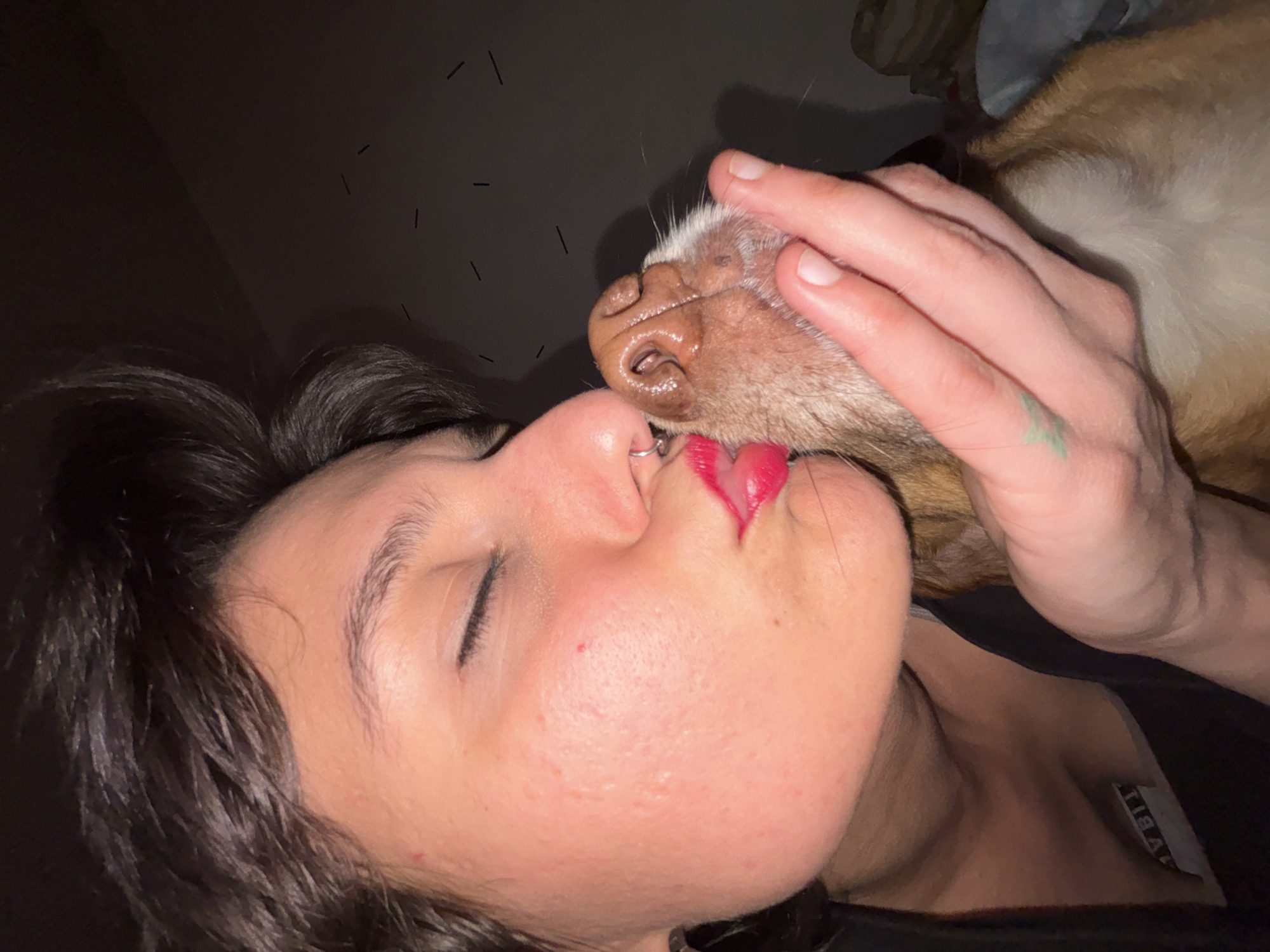 A person is kissing a dog. The person has dark hair and red lipstick and is wearing a nose ring. The dog is tan and white and the person is holding the dog's head. The background is dark. The location is not discernible and therefore cannot be identified as Sofia, Bulgaria.