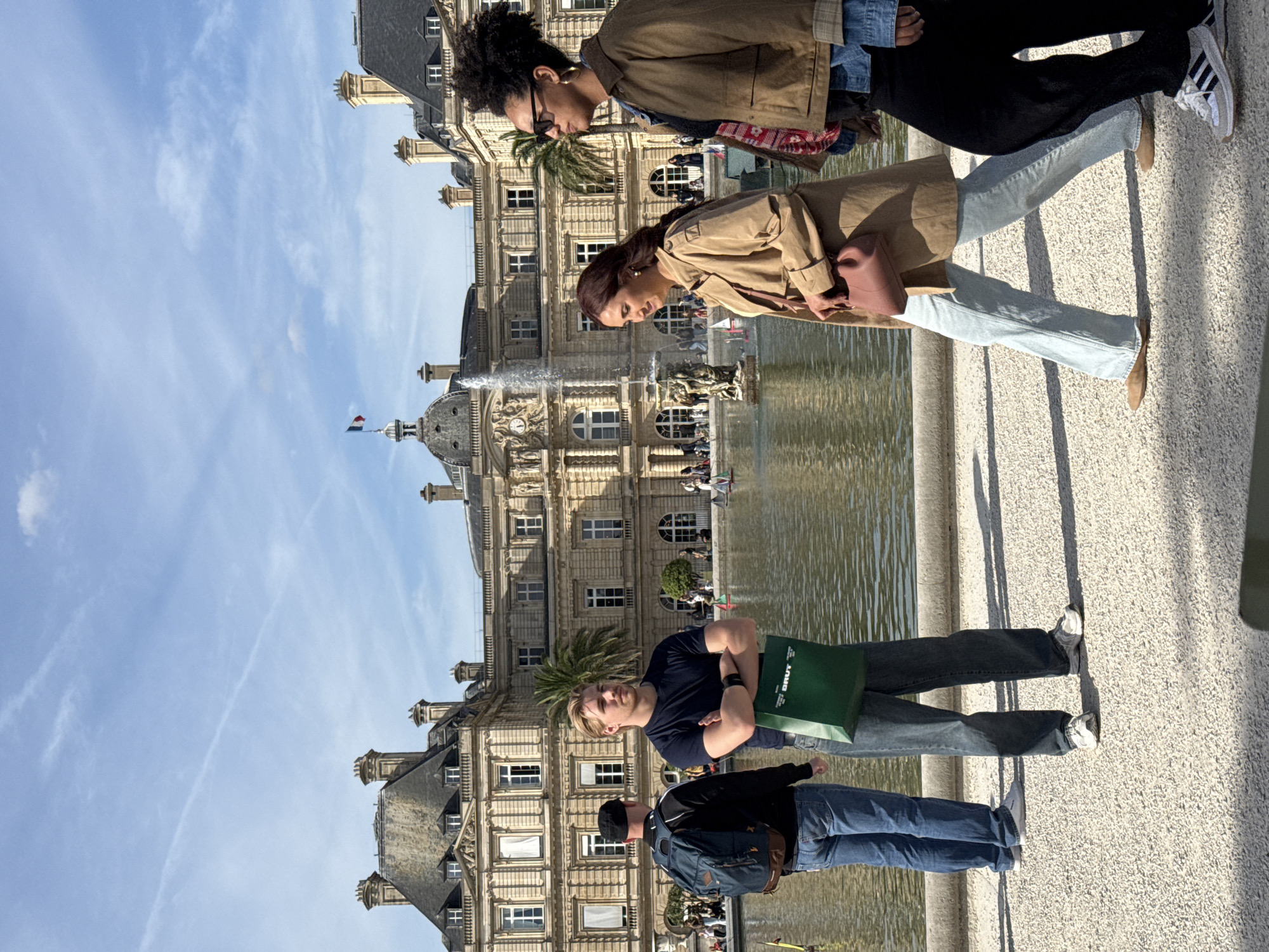 The photograph depicts a scene in the Jardin du Luxembourg in Paris, France. In the foreground, several people are standing on a stone pathway. On the left side, there are two men; one is wearing a black shirt, jeans, and is holding a green shopping bag, while the other is wearing a black jacket, blue jeans, and a backpack. On the right side, there are two women standing; the first woman is wearing a tan coat, blue jeans, and brown shoes, and the second woman is wearing a brown coat, black pants, and white sneakers. In the background is the Palais du Luxembourg, reflecting in the water of a large fountain. The sky is blue with wispy clouds. The lighting indicates it is daytime.