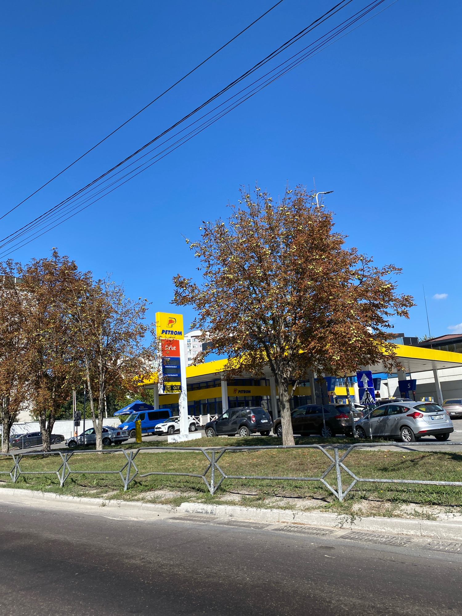 The image captures a street scene in Chișinău, Moldova, featuring a Petrom gas station. The gas station is identifiable by its prominent yellow and blue branding, with a tall sign displaying the Petrom logo and fuel prices. Several cars are parked at the station, including a blue van, a silver hatchback, and a dark SUV. Trees with autumnal foliage, exhibiting a mix of brown and some remaining green leaves, line the area in front of the gas station. Overhead power lines stretch across the clear blue sky. A metal fence separates the sidewalk from the grassy area. The foreground shows a paved road, likely a street or highway, with some wear and tear. The overall impression is a typical urban landscape on a sunny day.
