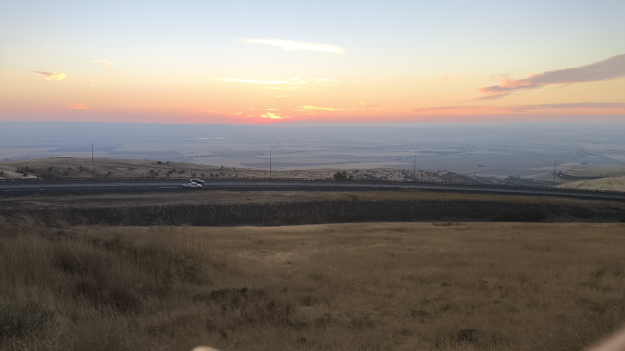 A wide-angle, high-angle shot captures a sunset over a vast, hazy valley near Tutuilla, United States. The sky is a gradient of pale blue at the top, transitioning through hues of orange and pink from the setting sun, which is partially obscured by a thin layer of clouds. A distant, flat horizon features a body of water and a sprawling landscape of fields, possibly farmland, with some visible structures. In the mid-ground, a highway curves across a gently sloping, dry, grassy hillside. Two cars are visible on the highway; a white car is in the right lane, and a black car is in the left lane, both moving away from the viewer. Streetlights are spaced along the highway. The foreground is dominated by a field of dry, golden grass, with some sparse, darker vegetation. The overall atmosphere is serene and expansive, with the warm tones of the sunset creating a peaceful mood.