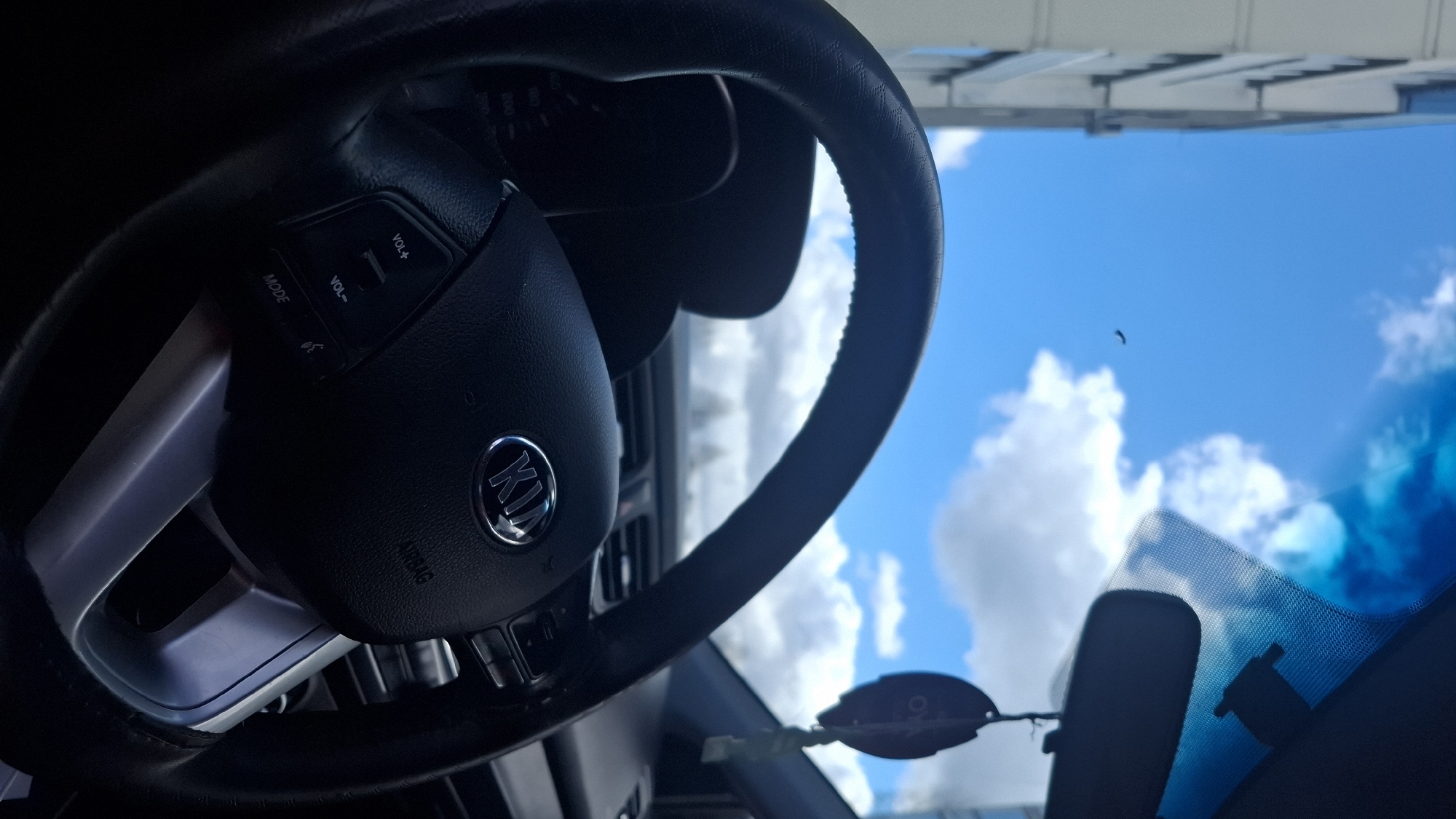 The image captures a view from inside a car, oriented vertically as if rotated 90 degrees counter-clockwise. The left side of the frame is dominated by a black steering wheel with silver-colored spokes. The center of the steering wheel features a prominent "KIA" logo within a chrome ring on the airbag cover, which also has the word "AIRBAG" embossed below. Control buttons are visible on the upper left spoke of the steering wheel, labeled "VOL+", "VOL-", "MODE," and an icon resembling a phone receiver for call management. The dashboard or air vents are dimly visible behind the steering wheel.
Through the car's windshield, the right side of the image reveals a bright blue sky dotted with scattered white, fluffy clouds, indicating clear and sunny weather conditions during daylight. A small, dark, crescent-shaped object is visible against the blue sky, possibly a bird in flight or a mark on the windshield. A rearview mirror is partially visible in the lower right, and a dark, leaf-shaped object, likely a hanging car air freshener or tag, dangles near it. Above the horizon line, along the top edge of the frame (which would be the right if correctly oriented), part of a building or structure with horizontal elements is discernible.
No people or specific activities are depicted. The scene is a static interior car shot looking outwards. The car is identifiable as a Kia. The provided context places this scene in Марковский, Russia.