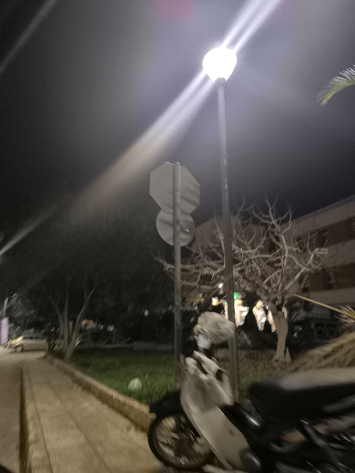 This image depicts an outdoor night scene in Drapetsona, Greece, characterized by artificial illumination. A tall street light pole dominates the mid-right, featuring a brightly glowing, spherical lamp that projects intense light rays and prominent lens flares across the dark sky. Positioned behind this, another pole holds two traffic signs, one octagonal above a circular one, but any text on them is illegible due to the angle and lighting conditions.
In the foreground on the right, a light-colored scooter or motorcycle is parked, with its front section, handlebars, and part of the seat visible. To the left, a paved sidewalk recedes into the distance, alongside a narrow strip of green grass. Various trees are scattered throughout the scene; some display bare, intricate branches against the dark background, suggesting a winter season or specific tree type, while others retain denser foliage.
A multi-story building is partially visible in the background on the right, with faint lights emanating from its interior. Further down the street on the left, a light-colored car is dimly discernible. The time of day is clearly night, with a dark, clear sky. The scene is static, showing no specific activity or event. No legible text is visible within the image. The overall mood is quiet and still, typical of an urban area after dark.