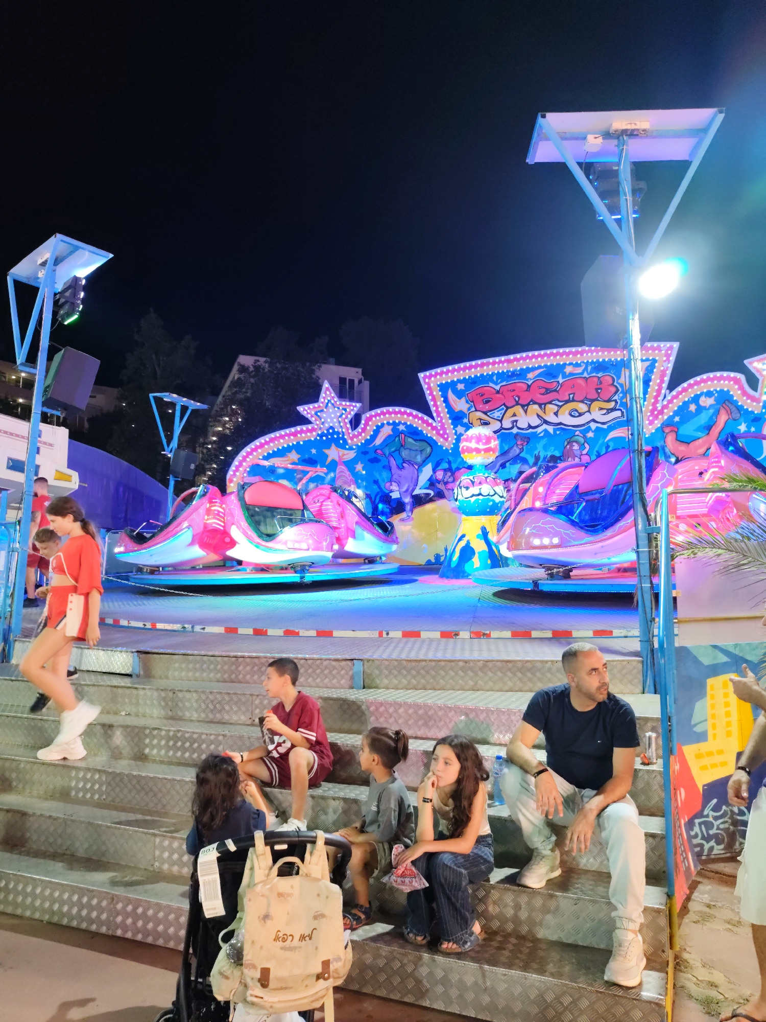 This image depicts an outdoor amusement park scene at night in Nesebar, Bulgaria, likely at the Sunny Beach resort. In the background, a brightly lit amusement ride, labeled "BREAK DANCE," is visible, featuring pink and blue lights and cartoonish figures. In the midground and foreground, several people are seated on metal bleacher-style steps.
To the left, a young woman in a red outfit walks down the steps. A young boy in a red shirt and shorts is seated on the steps, holding a small object. To his right, a young girl with long dark hair is also seated. Further to the right, a man in a dark t-shirt and light-colored pants is seated, looking off to the side. Another young girl in jeans and a white tank top sits next to him, with her hand on her chin. A baby stroller with a patterned bag is positioned on the steps in front of the seated children.
The environment is characterized by artificial lighting from the amusement ride and streetlights, creating a vibrant and somewhat surreal atmosphere. The steps appear to be made of diamond plate metal. Palm fronds are visible on the right side of the frame.