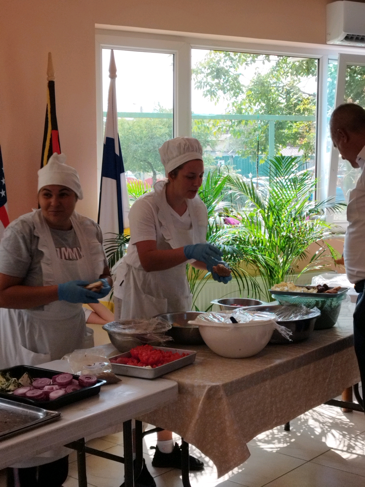 The image depicts an indoor scene, likely a community gathering or event, possibly in Bucharest, Romania, given the presence of what appears to be national flags. Two women, dressed in chef's hats and aprons, are preparing food at a table. The woman on the left is holding a small food item in her gloved hands, examining it. The woman on the right is also wearing gloves and appears to be working with food in a bowl. Various food ingredients are laid out on the table, including chopped red tomatoes in a rectangular tray, sliced red onions in another tray, and several bowls containing food, some covered with plastic wrap. In the background, a large window overlooks an outdoor area with trees and foliage, suggesting a sunny day. Two flags are visible, one with black, yellow, and red stripes and another with blue and white squares. The overall atmosphere appears to be one of food preparation and communal activity.