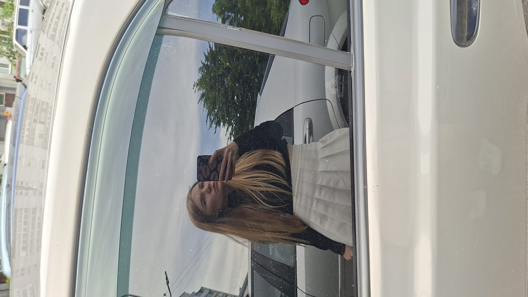 An image, taken outdoors in Chaikovsky, Russia, depicts a young woman with long blonde hair taking a selfie in the reflection of a car window. She is wearing a black turtleneck crop top and a white pleated skirt. Her left hand is holding a phone with a black case featuring a lips pattern, and she is wearing a ring on her right ring finger. The reflection also shows a silver car, green trees, and a cloudy sky. The car appears to be parked on a street with buildings visible in the background. The surface of the car window is speckled with dust.