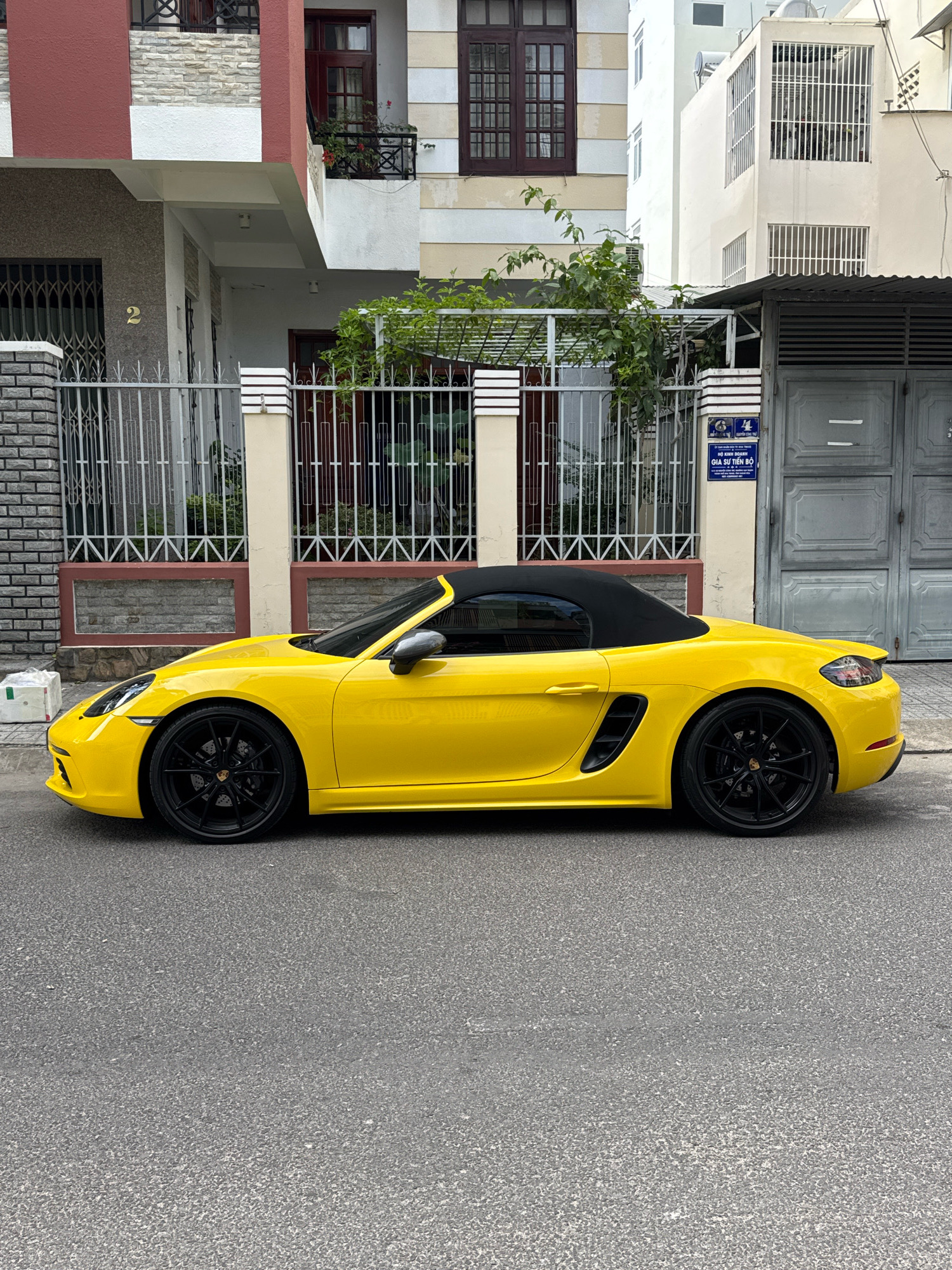 A bright yellow Porsche Boxster convertible is parked on the side of a paved street in Nha Trang, Vietnam. The car is positioned parallel to the curb, with its black soft top up. The vehicle features black alloy wheels and has a sleek, sporty design.
In the background, a modern building with a red and white facade is visible. The building has multiple windows, some with dark wooden frames and decorative metal grilles. A white balcony with plants is also present. To the right of this building, another white building with multiple windows and metal bars is seen. A gray metal gate is situated in front of the building, partially obscuring a large metal door. A sign with Vietnamese text is affixed to the wall next to the gate. The street is lined with a low fence and a brick pillar. The environment appears to be an urban residential area.