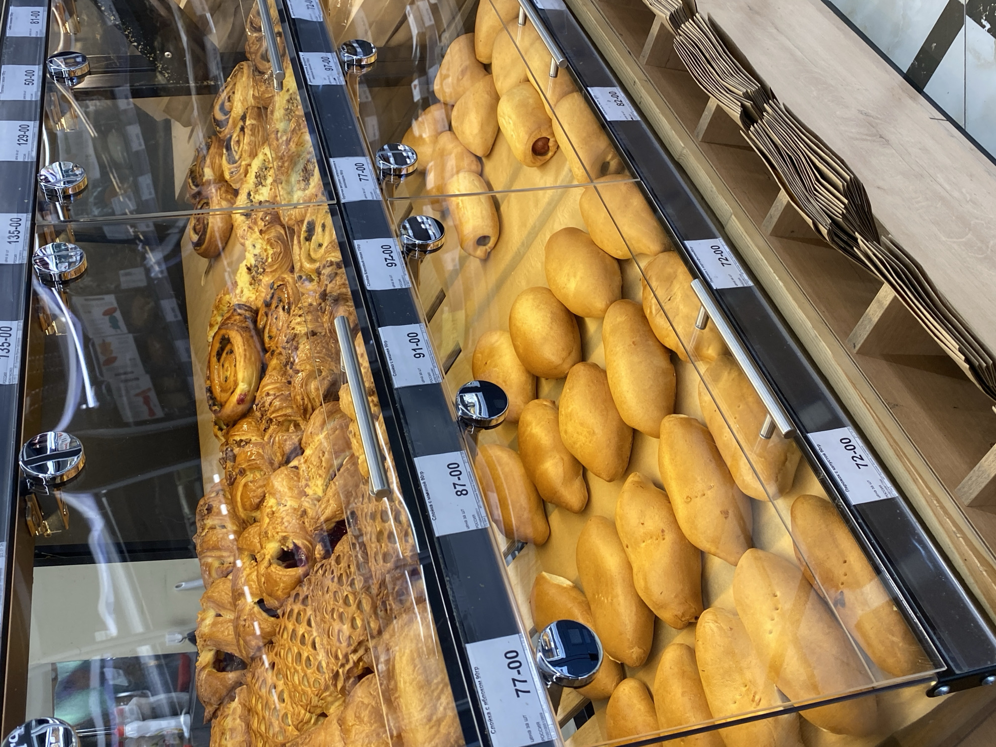 The image shows a bakery display case. The case contains three sections filled with pastries and bread. The section on the left features pastries with visible layers and various fillings. The central section contains filled buns, some with a visible sausage filling. The section on the right contains oblong bread rolls. The display case is marked with black price tags indicating the cost of each item in Russian rubles, including "77-00", "87-00", "91-00", "97-00", "72-00", "82-00", "135-00", "129-00", "50-00", and "81-00". The price tags also include the product name in Russian, like "Слойка с яблоком 90гр" (apple pastry 90g). The label indicates that the products originate from Russia. To the right of the bread rolls, there are stacks of brown paper bags.