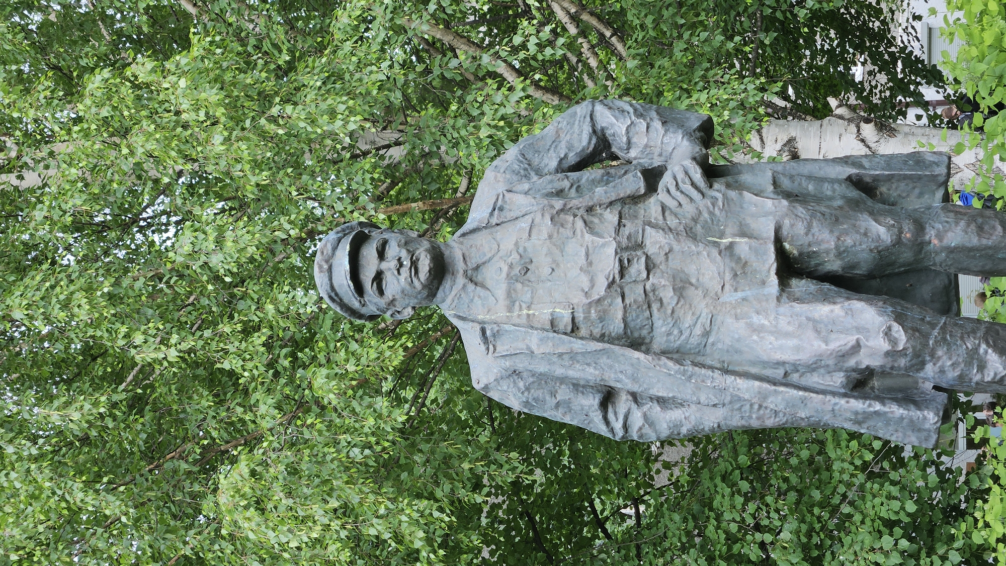 The image shows a bronze statue of a man wearing a cap and a long coat, set against a backdrop of lush green trees. The statue is positioned at an angle, with the man's head and upper body facing towards the left of the frame. His face is rendered with some detail, showing a stern expression. The texture of the bronze is rough and somewhat weathered, with hints of verdigris. The man's clothing appears to be a military-style uniform, judging by the structure of the coat and the cap.
The background is dominated by the dense foliage of trees, with branches and leaves filling much of the frame. Some of the branches are bare, suggesting a possible contrast with the fullness of the leaves. In the upper right corner, a portion of a white building with windows is visible, partially obscured by the trees, hinting at an outdoor, urban or park-like setting. There are a few indistinct figures visible in the background, seemingly observing the statue, adding a sense of scale and context. The lighting suggests it is daytime, with no strong shadows indicating a specific time. The overall impression is one of a public monument set within a natural environment.