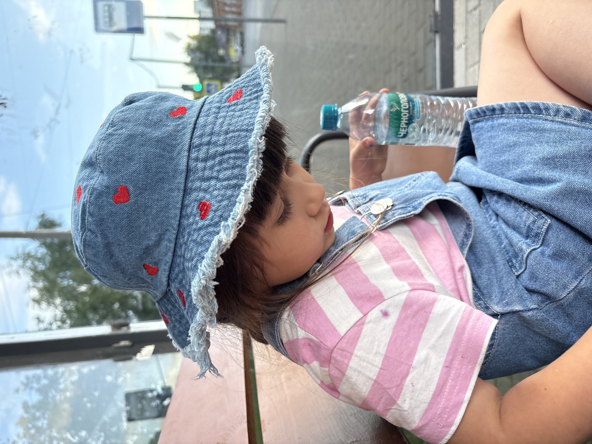A young child is seated on a reddish-brown bench, observed from a slightly elevated perspective, looking downward. The child wears a blue denim bucket hat embroidered with red hearts and featuring a frayed brim. Underneath the hat, brown hair is visible. The child's attire includes a pink and white horizontally striped t-shirt, which exhibits faint purple stains on the left chest area, and blue denim overalls. A silver-colored chain with circular charms is draped across the left side of the child's chest. In their right hand, the child holds a transparent plastic water bottle with a blue screw cap. The bottle's label features the text "ЧЕРНОГОЛОВ" (Chernogolovka), identifying it as a Russian brand. Behind the child, through a glass panel, an outdoor urban scene is visible, including a blurred green traffic light, a distinct bus stop sign (white with a blue border and bus icon), paved ground, and indistinct background foliage and structures under a partly cloudy sky. The setting is consistent with a bus stop in an urban environment in Russia, specifically within the city of Chaikovsky.