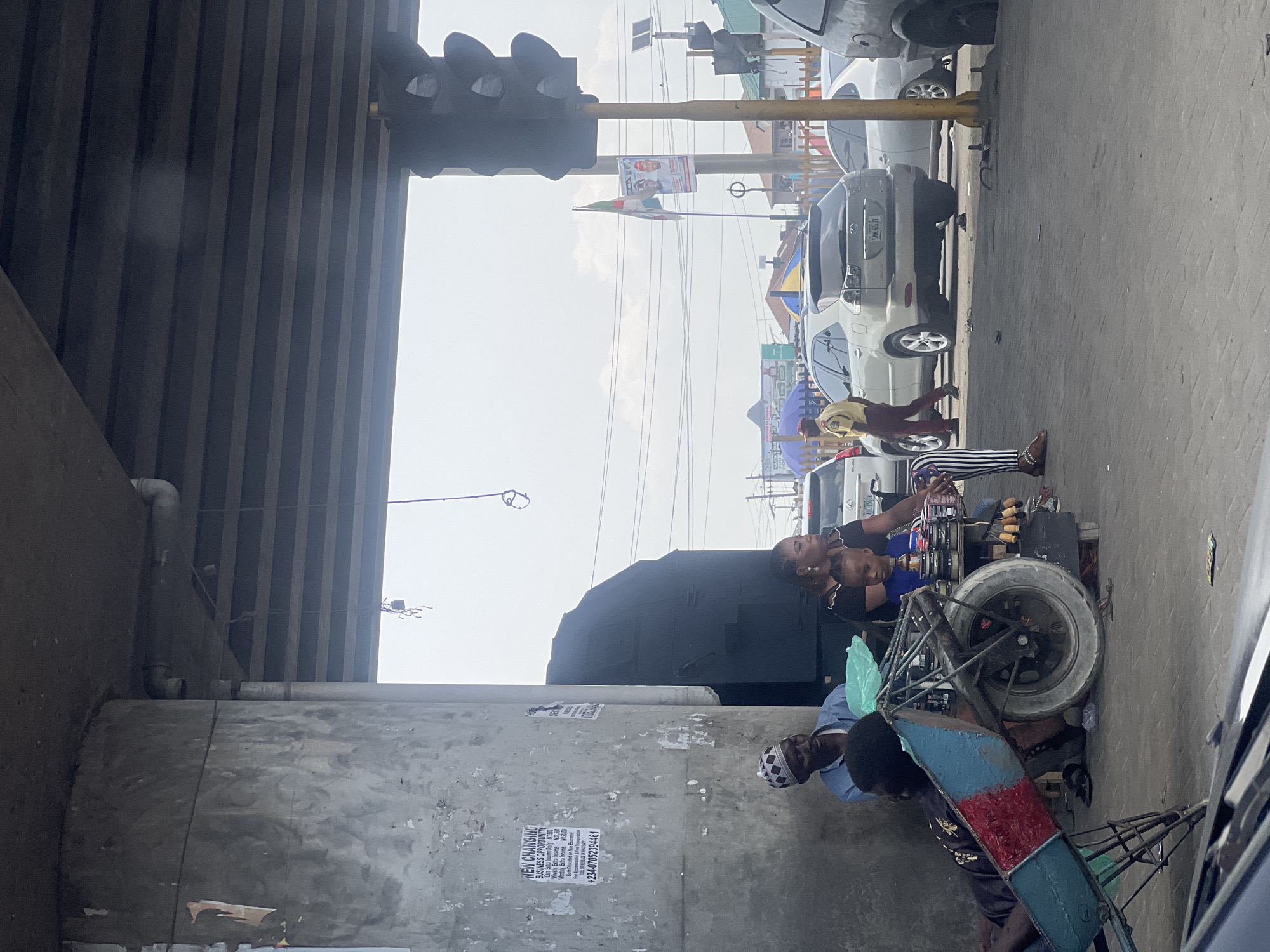 Here is a forensic description of the image:
The image, taken in Ado, Nigeria, shows a street scene under a highway overpass during daylight. A utility pole is partially visible. In the foreground, a light-skinned person in a white and black patterned head covering is seated on the ground next to a vendor's cart. The cart includes a blue and red container/cabinet mounted on a wheeled frame. Two other individuals are positioned on the cart. A light-skinned individual wearing a blue shirt is seated on the cart, behind a dark-skinned individual wearing a black shirt. Traffic and commercial buildings are visible in the background.