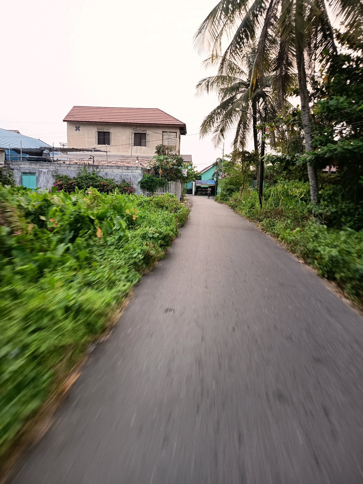 The image depicts a straight, paved road leading into the distance, flanked by lush greenery on the left and a line of palm trees and dense vegetation on the right. In the background, a two-story building with a reddish-brown tiled roof and cream-colored walls stands prominently on the left. A smaller building with a blue roof is visible to its left. Further down the road, on the right side, a light blue building with a sign reading "SMP NEGERI 1 BANJARBARU" is situated. A solitary person is walking away from the camera on this road. The sky is overcast, suggesting a cloudy day. The motion blur indicates the photo was taken while moving.