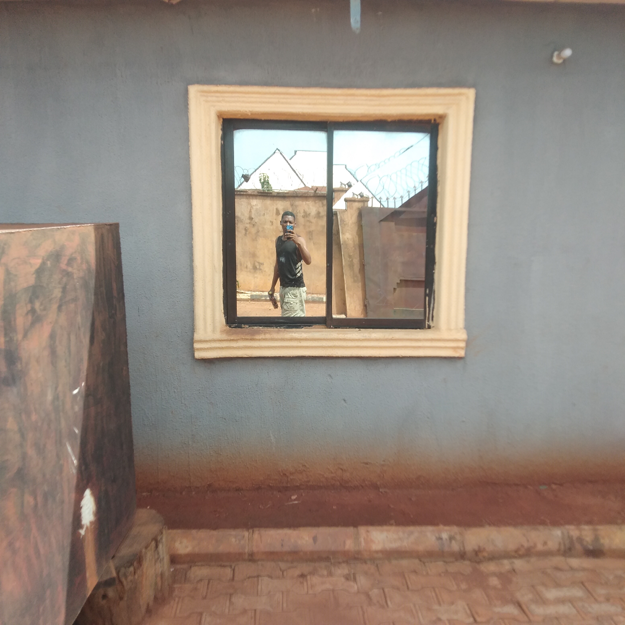 A medium shot depicts a building exterior in Unknown, Unknown. The wall is painted gray with a wide, brown line near the base. A tan-trimmed window with a black frame reflects a man holding a phone. Outside, the reflection also includes a tan wall topped with barbed wire. To the left of the window is a brown object. The ground is paved with interlocking bricks.
