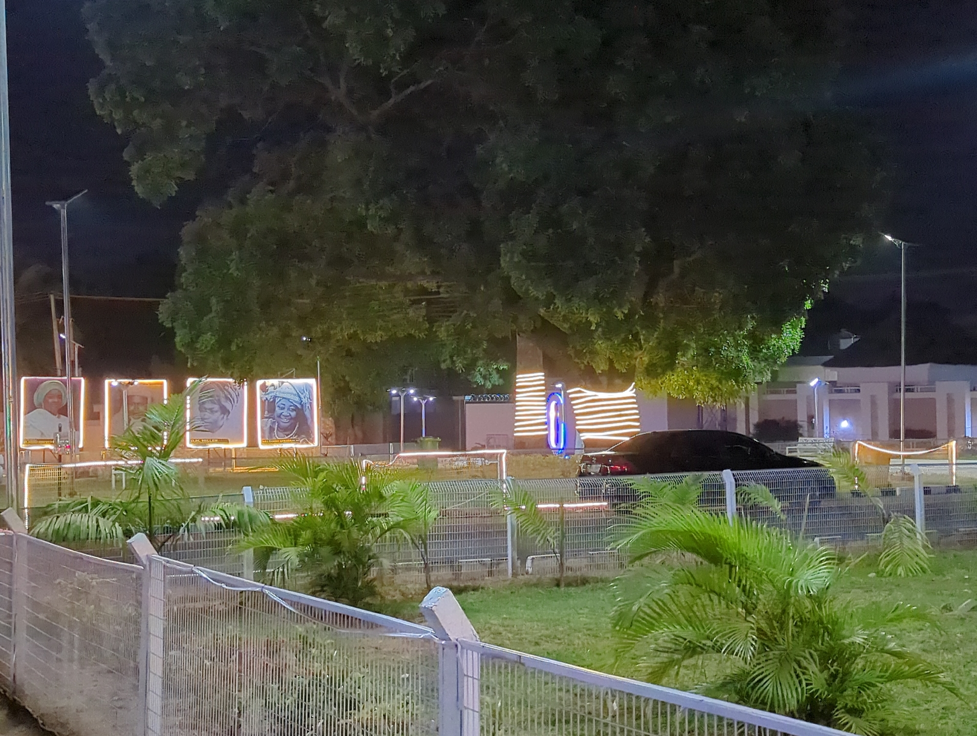 Observed at night in what is purportedly Unguwar Rimi, Nigeria, the scene captures a fenced area, possibly a park or memorial. Four illuminated portraits are visible, depicting individuals, possibly notable figures. String lights delineate the perimeter of the area. A large tree dominates the upper portion of the frame. A dark-colored vehicle is partially visible behind some of the displays. Streetlights are present along the path in the distance. Palm-like plants are scattered throughout the enclosed area. A gray metal fence is in the foreground. The area appears to be illuminated by artificial lighting.