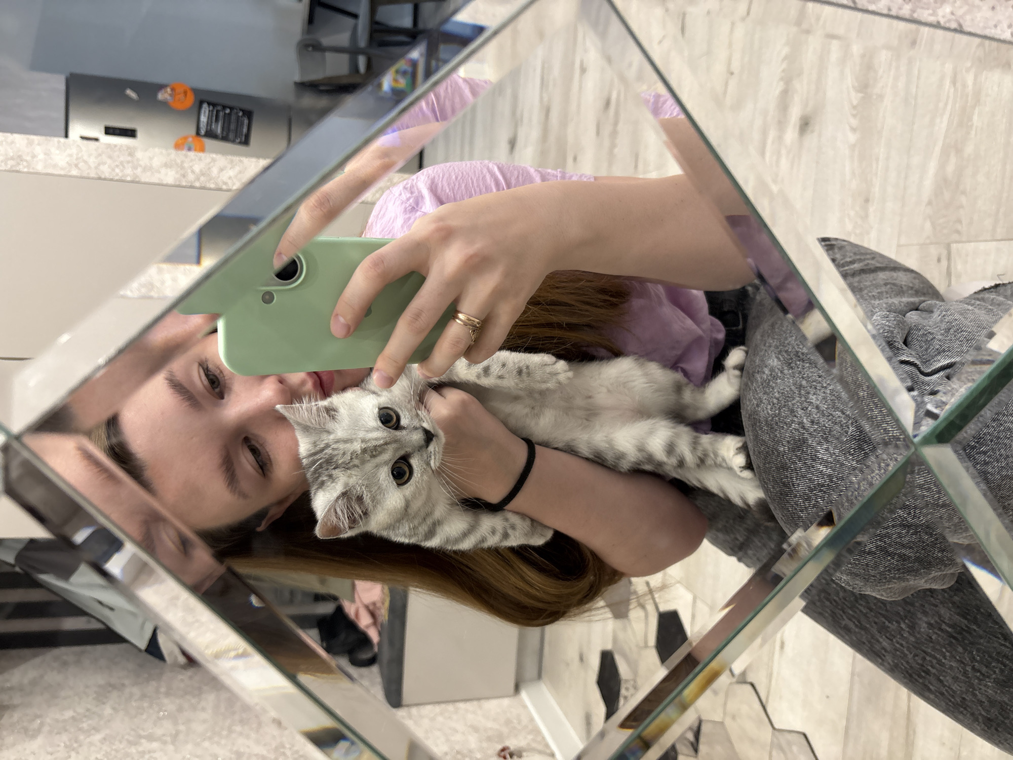 A young woman takes a mirror selfie with her small cat in an indoor setting, likely a home in Izhevsk, Russia. The image is captured through a faceted mirror, creating multiple reflections and a fragmented view of the scene.
The woman is positioned towards the right side of the frame. Her head is tilted, and she has light brown hair, visible eyebrows, and a calm expression with a hint of a smile. She is wearing a light purple top. Her left arm gently cradles a small, light grey tabby cat with prominent black stripes and large, round amber eyes. The cat is looking directly at the camera with an attentive and curious gaze. A black hair tie is worn on the woman's left wrist. Her right hand holds a light green smartphone, which she is using to capture the image. A gold-colored ring with a small charm is visible on one of her fingers.
The background, seen through various reflections in the mirror, reveals elements of a domestic space. A light-colored countertop or table is visible, along with a silver refrigerator adorned with small orange magnets or decorations. The floor appears to be made of light-colored wooden planks or hexagonal tiles. Parts of the woman's lower body, dressed in grey textured fabric, are also visible through the mirror's reflections. The overall atmosphere is casual and affectionate, highlighting the bond between the woman and her pet. No visible text is present in the image.
