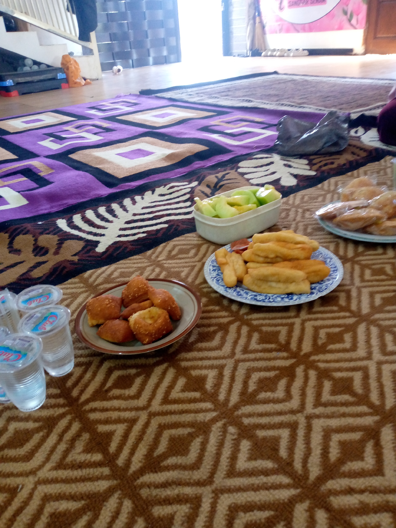 The image shows a close-up of food items laid out on a patterned carpet, likely in a domestic setting within the city of Mojokerto, Indonesia. In the foreground, there are several plastic cups filled with clear liquid, possibly water. To the right of the cups, a plate holds an assortment of fried snacks, appearing to be dough-based and golden brown. Further to the right, a white container holds cut pieces of fruit, primarily green, which could be melon or cucumber. Beyond these, another plate is visible, containing more fried pastries or bread items, some of which are still in plastic packaging. The carpet itself has a geometric pattern in brown and beige tones. In the background, a purple and black patterned rug is partially visible, and further back, stairs and a doorway can be seen, suggesting an indoor environment. The lighting suggests it might be daytime.