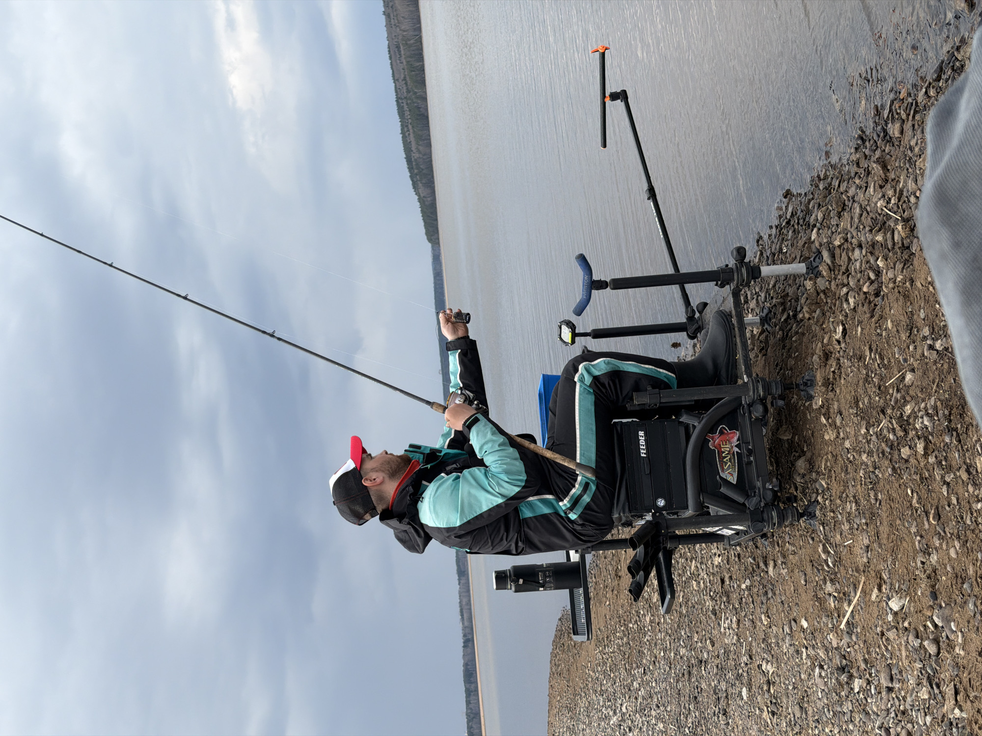 A man is fishing on the shore of a lake or river near the city of Chaikovsky, Russia. He is seated in a specialized fishing chair equipped with rod holders and other accessories. He wears a black and blue jacket, black pants with a light blue stripe, and a red and black baseball cap. He holds a fishing rod in his hands, appearing to be reeling in his line. The shoreline is composed of gravel and dirt. The water stretches into the distance, with a forested area visible on the opposite shore. The sky is overcast. The image is rotated 90 degrees counter-clockwise.