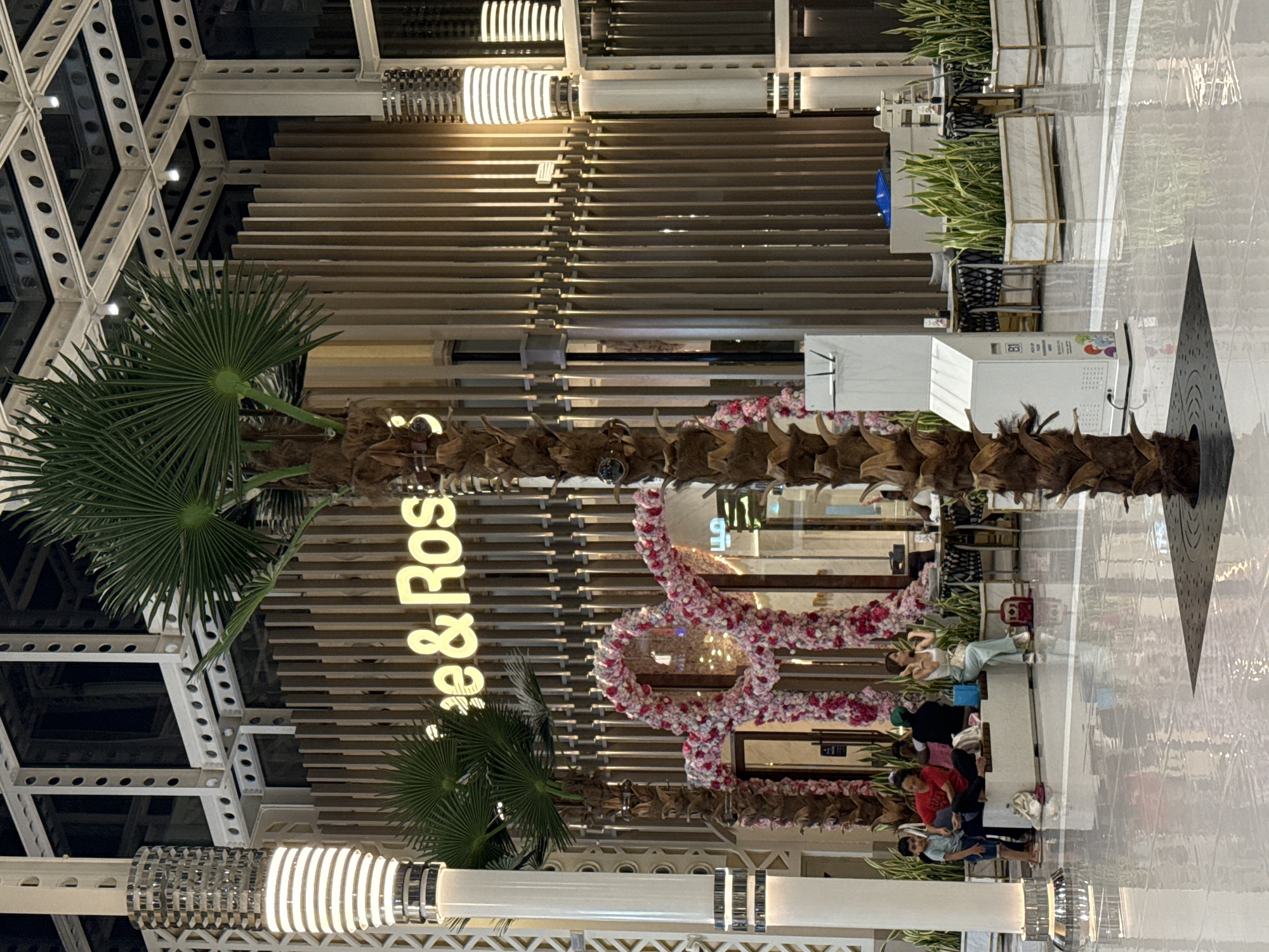 This image depicts the interior of a modern shopping mall in Dubai, United Arab Emirates. In the foreground, a large, prominent palm tree stands centrally, its textured trunk and fronds contrasting with the polished, reflective floor. To the left and right, tall, cylindrical light fixtures with ribbed detailing illuminate the space.
Behind the palm tree, a storefront with a wooden slatted facade is visible, featuring a neon sign that reads "e & Ros". A heart-shaped floral archway, adorned with pink and white flowers, frames the entrance to the establishment. In front of the storefront, a white bench is occupied by several individuals. A young boy in a blue shirt and shorts stands to the far left, while a woman in a red top sits next to him. Further down the bench, another woman in a white top and light blue pants is seated, appearing to adjust her hair. Several bags are placed on the ground near the bench.
The environment is well-lit, with architectural elements of exposed metal beams and columns visible in the upper part of the frame, suggesting a contemporary and open design. Several potted plants in white planters add greenery to the scene. A white information kiosk with text and graphics is positioned to the left of the main palm tree. Tables and chairs are arranged in the background, indicating a seating area for patrons. The overall impression is of a clean, well-maintained, and possibly upscale retail or dining environment within a Dubai mall.