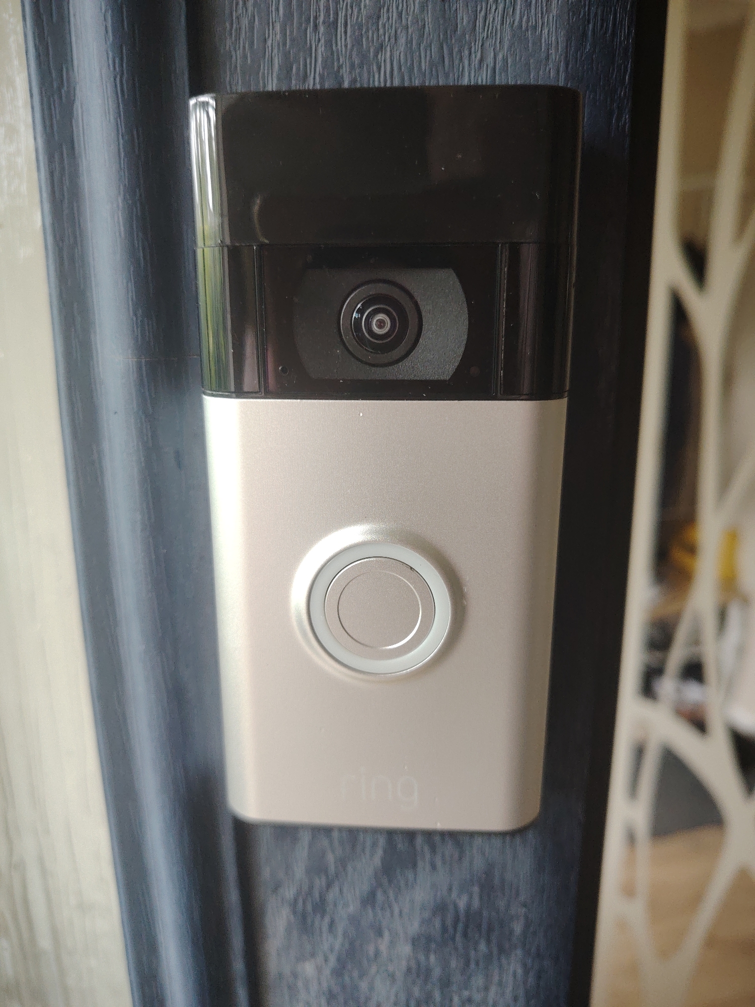 A Ring doorbell is mounted on a dark blue textured door. The doorbell is a silver and black device with a camera at the top and a circular button below it. The Ring logo is visible on the lower portion of the doorbell. To the left of the door, a dark blue curtain is visible. To the right of the door, a white decorative screen is partially visible. The image appears to be taken at eye-level. The location is the city of Huntingdonshire, United Kingdom.