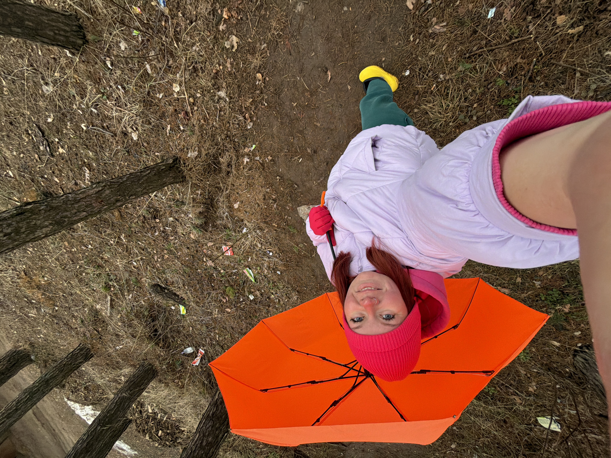 A ground-level, high-angle shot depicts a person holding an orange umbrella. The person is wearing a pink knit hat, a lavender jacket with a pink collar, green pants, and a yellow rain boot. The location appears to be a wooded area possibly in the city of Chaikovsky, Russia, as requested. The ground is covered with dry leaves, twigs, and some trash. Several tree trunks are visible. The person is looking up at the camera with a smile.