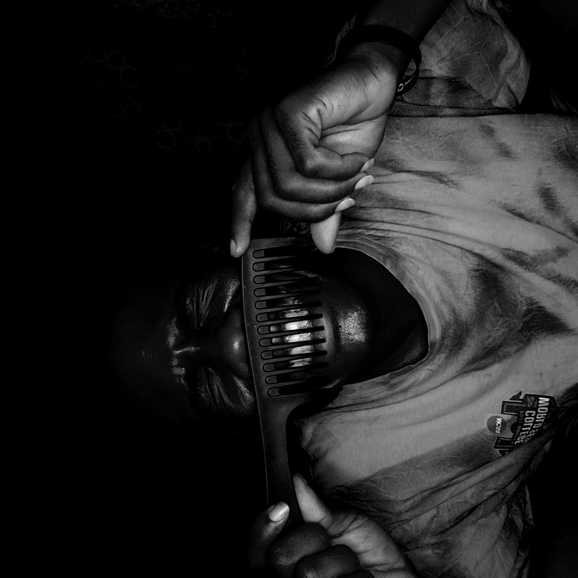 This black and white portrait, taken in Narayi, Nigeria, features a person positioned diagonally, with their head to the left and body extending towards the lower right. The scene is dimly lit, creating deep shadows that make the background almost entirely black, emphasizing the subject.
The central activity involves the person holding a wide-tooth comb (an afro pick) horizontally across their face, specifically over their mouth and lower nose. Their hands are prominent, with fingers curled around the comb, holding it in place. Through the teeth of the comb, the person's mouth is slightly open, revealing several teeth which stand out brightly against their dark skin and the dark comb.
The subject wears a light-colored, possibly tie-dye or patterned t-shirt. On the right side of the shirt (from the viewer's perspective), a logo or print is visible. The text on this logo appears inverted or reversed, reading "MIDWAY COLLEGE" and "ICT". A wristband is also visible on the person's right wrist. The person's facial expression, partially obscured by the comb, appears contemplative or ambiguous. The high contrast and the unusual placement of the comb give the image a striking and introspective quality.