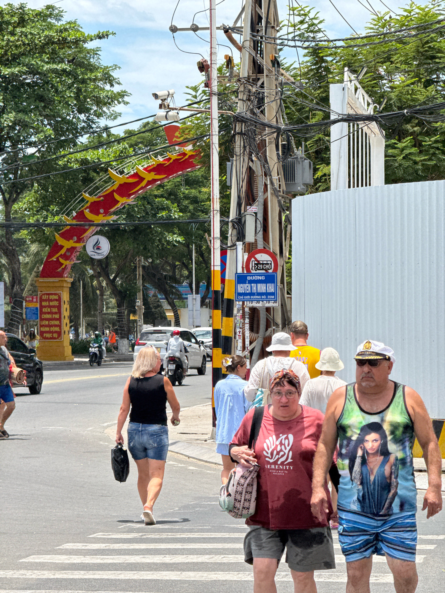 The image depicts a street scene in Nha Trang, Vietnam, characterized by a wide road with traffic, pedestrian activity, and overhead electrical infrastructure. In the foreground, four individuals are walking across a crosswalk. A woman in a black tank top and denim shorts carries a black bag. Beside her, a man in a green tank top with a woman's portrait on it and blue striped shorts walks forward. A woman in a red t-shirt and grey shorts is to the left of the man, carrying a patterned bag. Further left, partially obscured, is a person wearing blue shorts and carrying a rolled-up mat.
In the middle ground, traffic flows along the asphalt road. A black car is visible on the left, and several motorcycles and cars are further down the street. An arched gateway with red and yellow dragon motifs spans the road on the left side of the image, with a sign beneath it that includes Vietnamese text. Overhead, a complex network of electrical wires and poles dominates the upper portion of the frame, with a security camera mounted on one of the poles. To the right, a white corrugated metal fence and a partially constructed white structure are visible. The environment is outdoors, with lush green trees lining the street and a bright, possibly overcast sky.