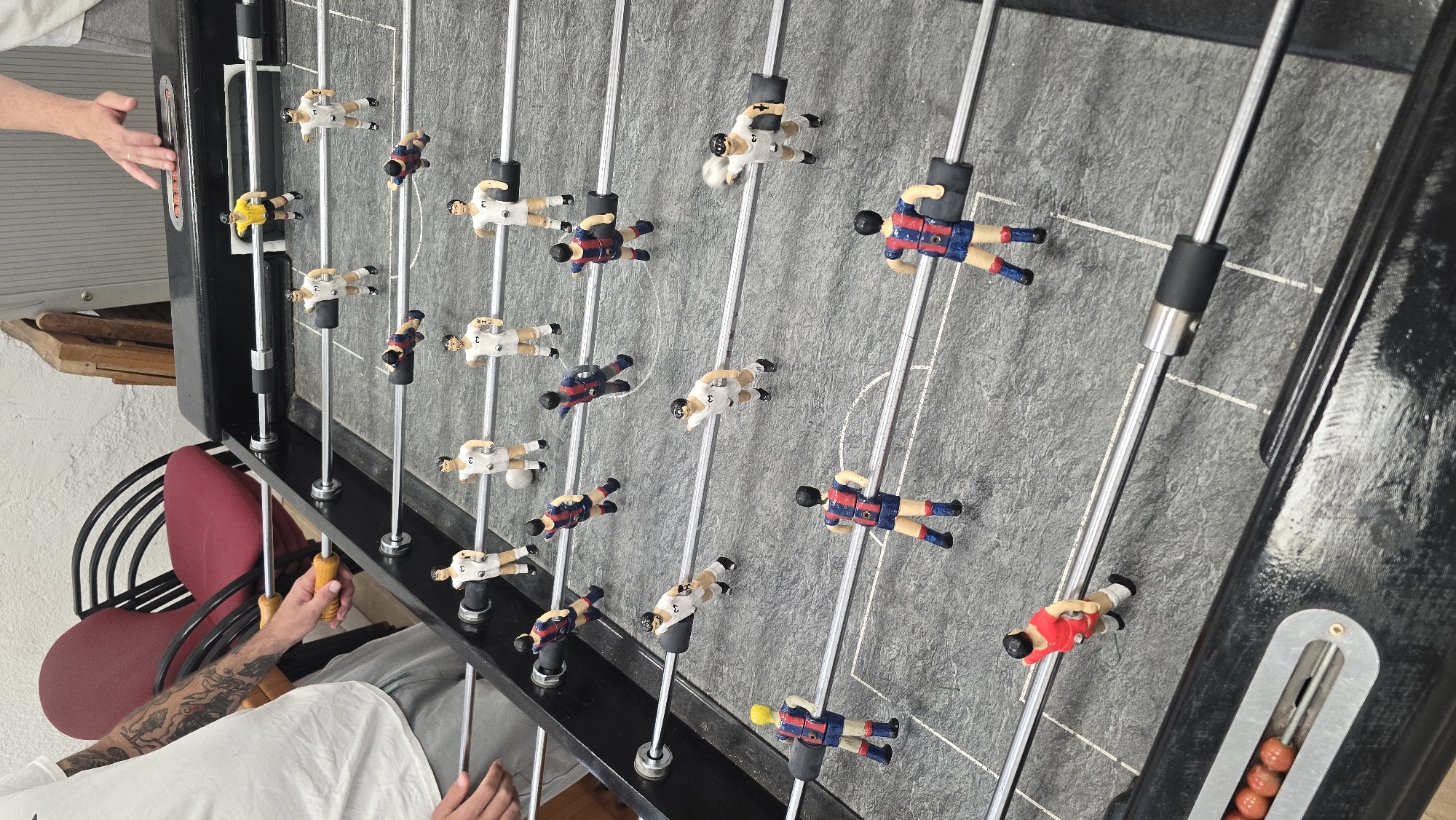 The image captures a top-down view of a foosball table, rotated 90 degrees counter-clockwise. The playing surface is a textured grey with white boundary lines and a central circle. Two teams of plastic players are visible, mounted on metal rods. One team wears white jerseys, some of which display the number "3" or the text "CHE". The opposing team wears red and blue striped jerseys, strongly resembling FC Barcelona's colors, which aligns with the given location context of Barcelona, Spain. There's also one player in a yellow jersey and another in a solid red jersey.
Several human hands are present in the frame, suggesting active play. On the left side, a pair of light-skinned hands is positioned near the edge of the table. On the bottom right, a person's forearm, adorned with tattoos, is visible, operating one of the foosball rods via a yellow wooden handle. This person is wearing a white t-shirt.
In the background, a red office-style chair with a black frame is prominent. Further back, elements of a room are visible, including a light-colored, textured wall and some wooden planks. One end of the foosball table shows a scoring mechanism with red beads. The overall scene depicts a casual game of foosball, likely indoors, with players engaged in the activity.