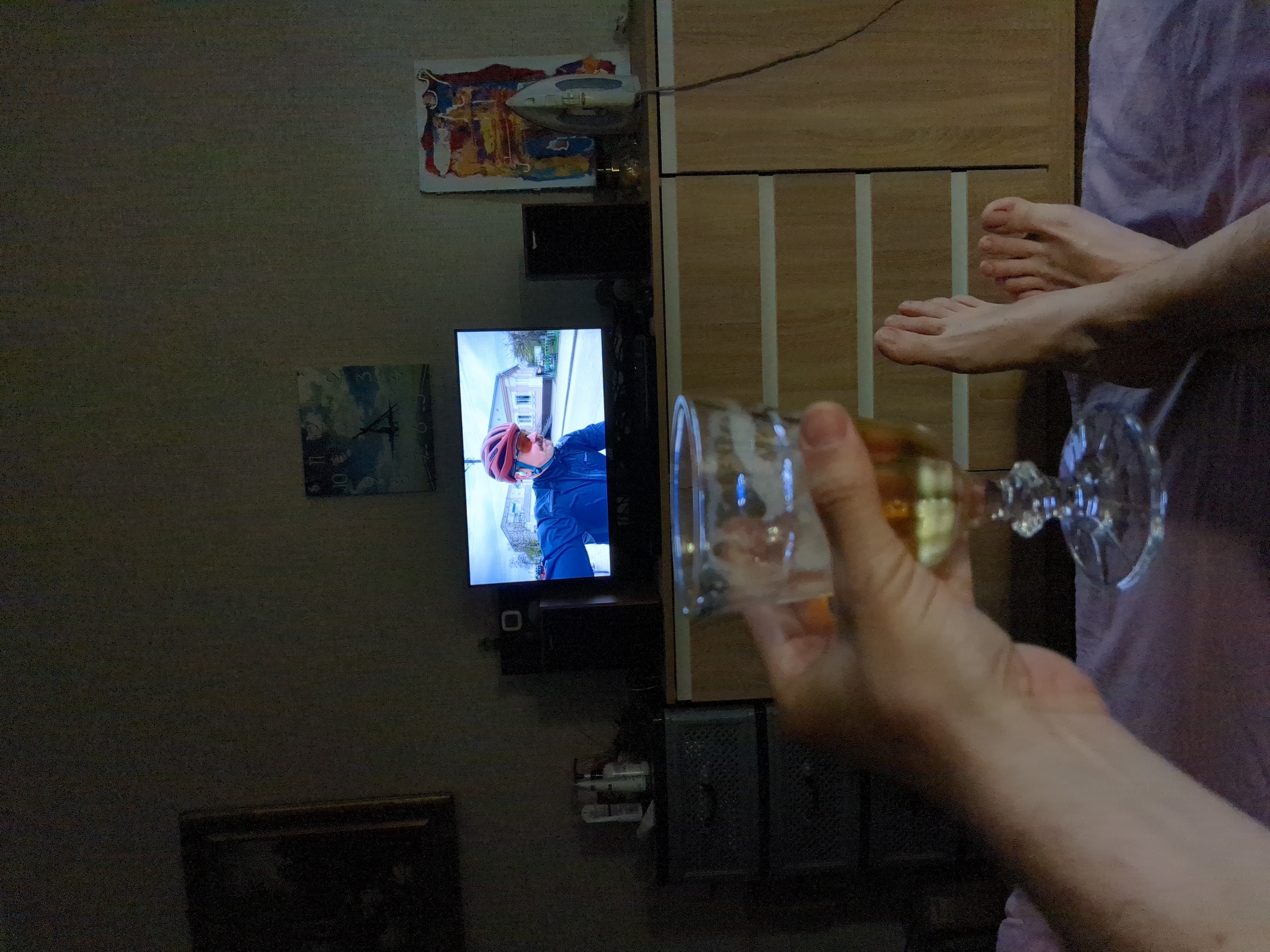The image depicts a dimly lit indoor scene. In the foreground, a person's left hand holds a stemmed glass filled with a pale yellow liquid, possibly alcoholic. The hand is positioned close to the camera, obscuring part of the view. To the right of the hand, a pair of bare feet are visible, resting on a purple surface, likely a blanket or bed.
In the mid-ground, a television screen displays an image of a man wearing a red helmet and glasses, riding a bicycle. The man is dressed in a blue jacket. The television is placed on top of a wooden dresser with multiple drawers. To the left of the television, there is a black speaker. On the dresser, to the right of the television, an iron is positioned next to a colorful abstract artwork. Behind the television, on the wall, a framed picture is hung, and another poster-like image with clouds and a silhouette of a plane is visible.
To the left of the television, on a dark gray plastic set of drawers, there are several bottles, likely toiletries. The overall environment appears to be a bedroom or living area, with muted colors and low lighting. There is no specific indication of the city of Boryspil, Ukraine, within the visual elements of the image.