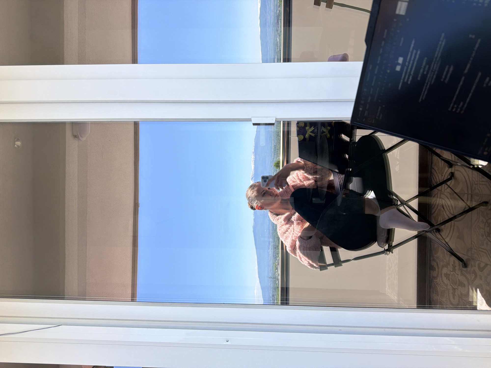 A woman is reflected in a window, holding a smartphone and wearing a pink sweater and black pants. She is seated on a black metal chair. The reflection shows a clear blue sky and distant mountains, suggesting an outdoor setting. A portion of a computer screen is visible in the upper right corner. The location is identified as Buhovo, Bulgaria, based on the available data. The interior space adjacent to the window appears to be a room with a window to the left. The floor is decorated with a carpet.